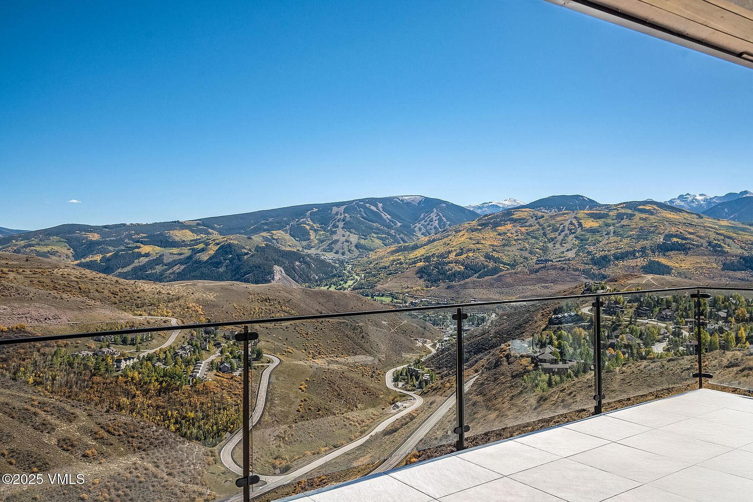 This image showcases a stunning view from a patio or balcony, featuring a modern glass railing and tiled flooring. The panoramic vista includes rolling hills, mountains, and a winding road, creating a sense of serenity and luxury. The clear blue sky enhances the overall appeal, making it an ideal outdoor space for relaxation and entertainment.