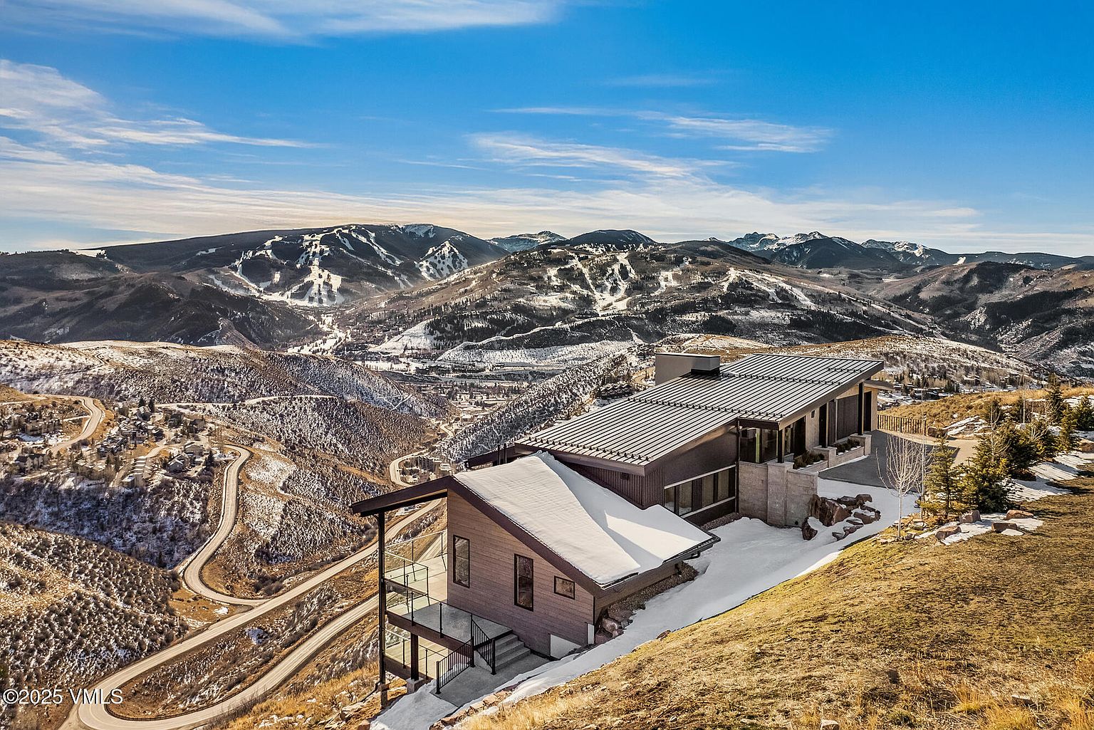 This aerial view showcases a modern mountain home nestled on a hillside, with stunning panoramic views of snow-capped mountains and winding roads. The house features a contemporary design with a metal roof, clean lines, and multiple levels, blending seamlessly with the natural landscape. The surrounding terrain is a mix of grassy slopes and patches of snow, emphasizing the property's unique location and access to outdoor recreation.
