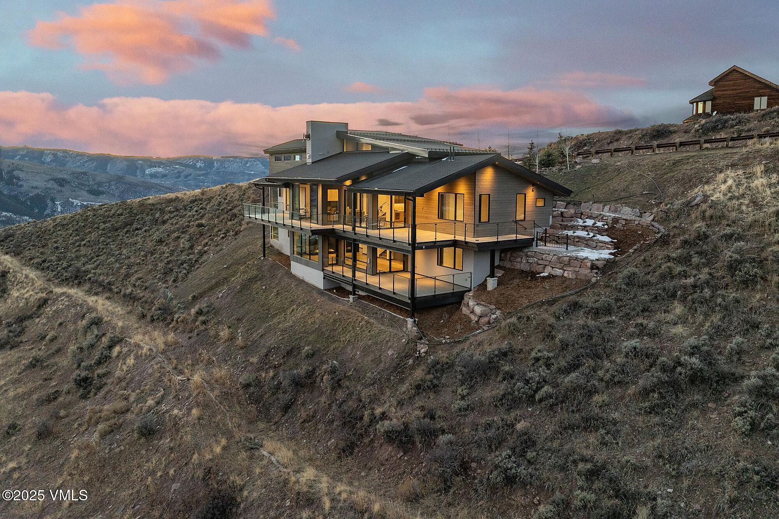 This aerial view showcases a modern, multi-level home nestled on a hillside. The house features a dark roof, light-colored siding, and expansive glass balconies offering stunning views. The surrounding landscape is a mix of natural terrain and carefully designed stone retaining walls, enhancing the property's appeal.