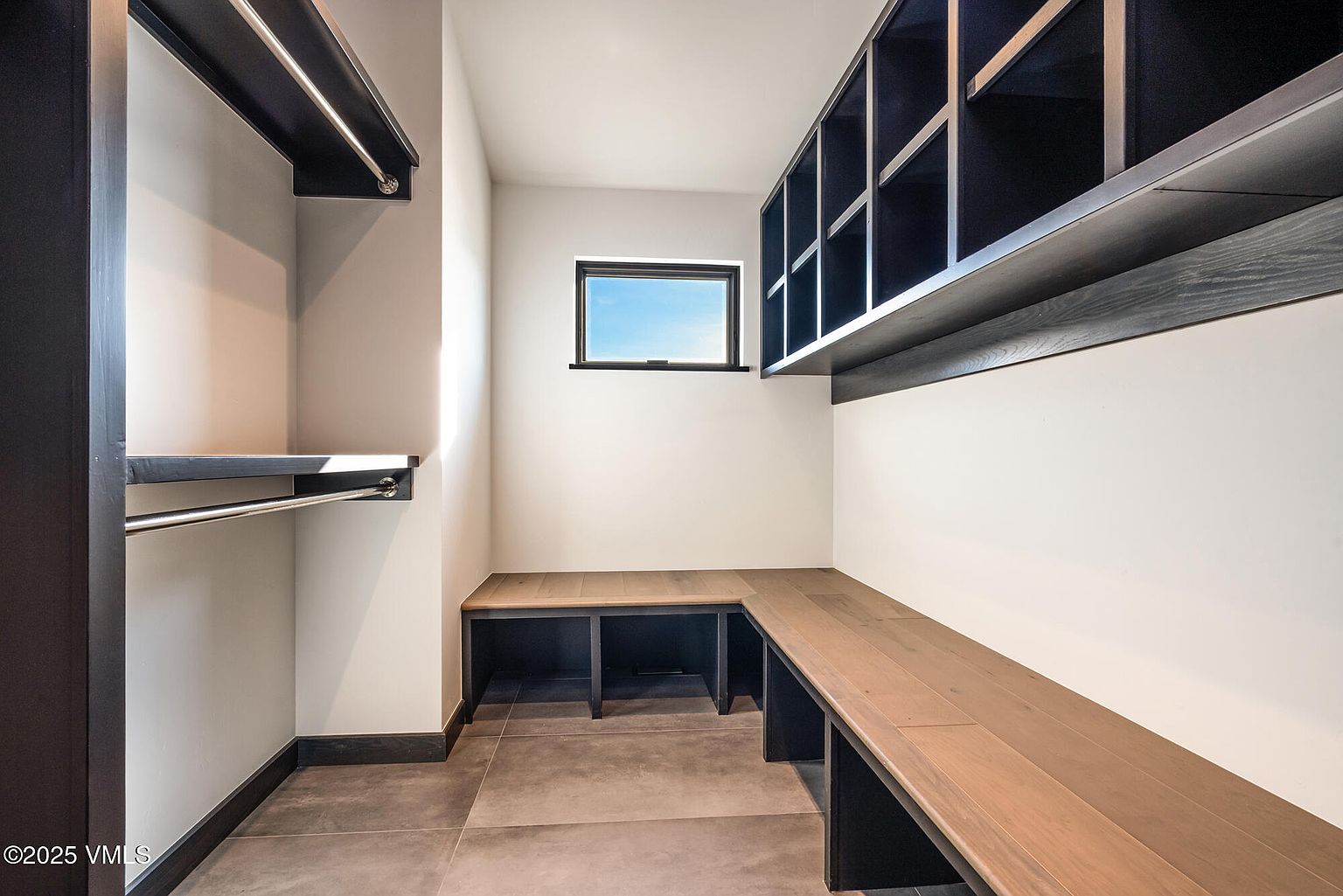 This is a well-organized walk-in closet featuring custom shelving and hanging rods. The closet includes a built-in bench with storage underneath and additional overhead storage cabinets. The color palette is neutral, with dark wood accents providing a modern and sophisticated look.