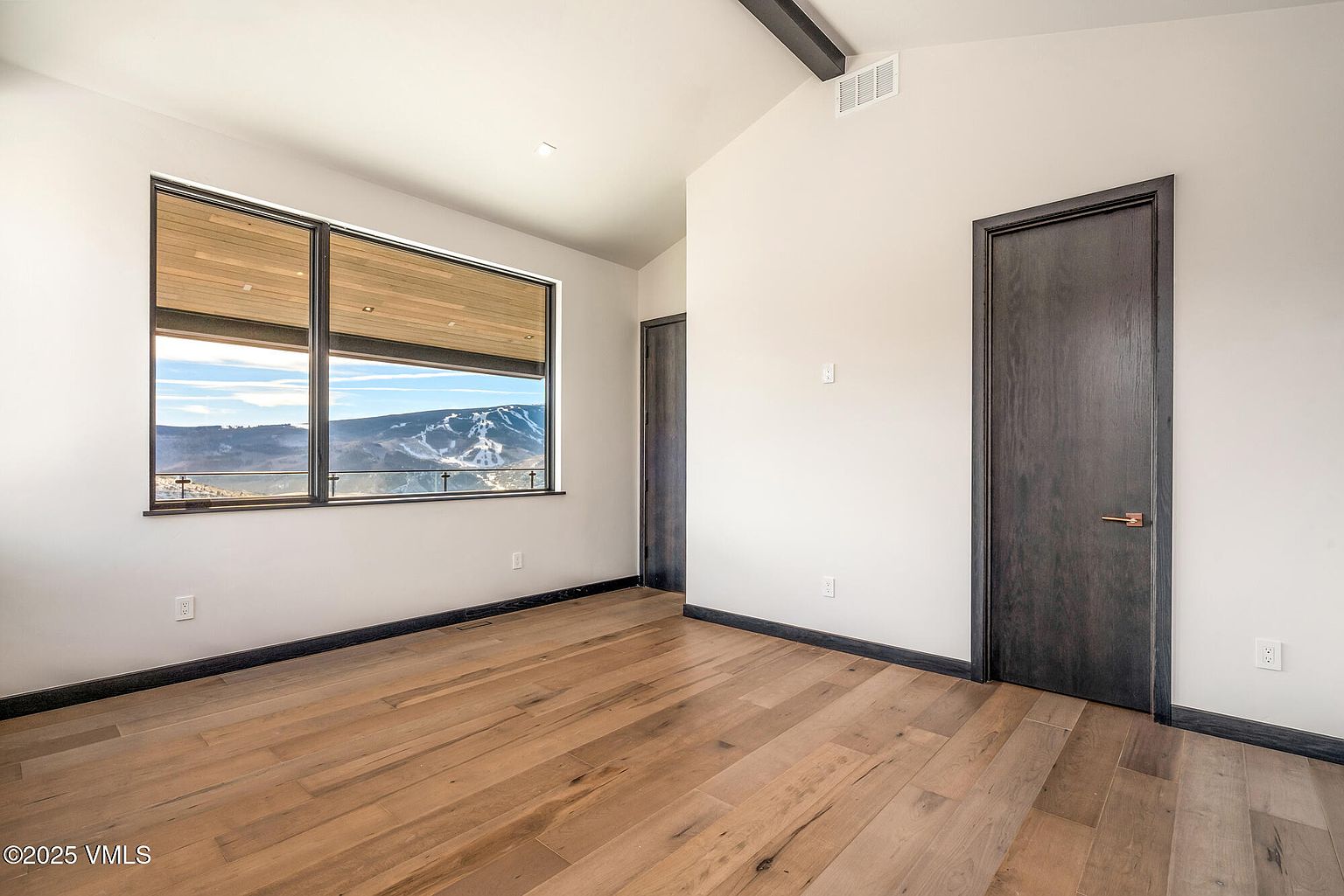 This is a primary bedroom featuring hardwood floors, neutral-colored walls, and a large window with a view of a mountain landscape. The room has modern doors with dark trim and a minimalist aesthetic. The space is bright and airy, creating a serene and inviting atmosphere.