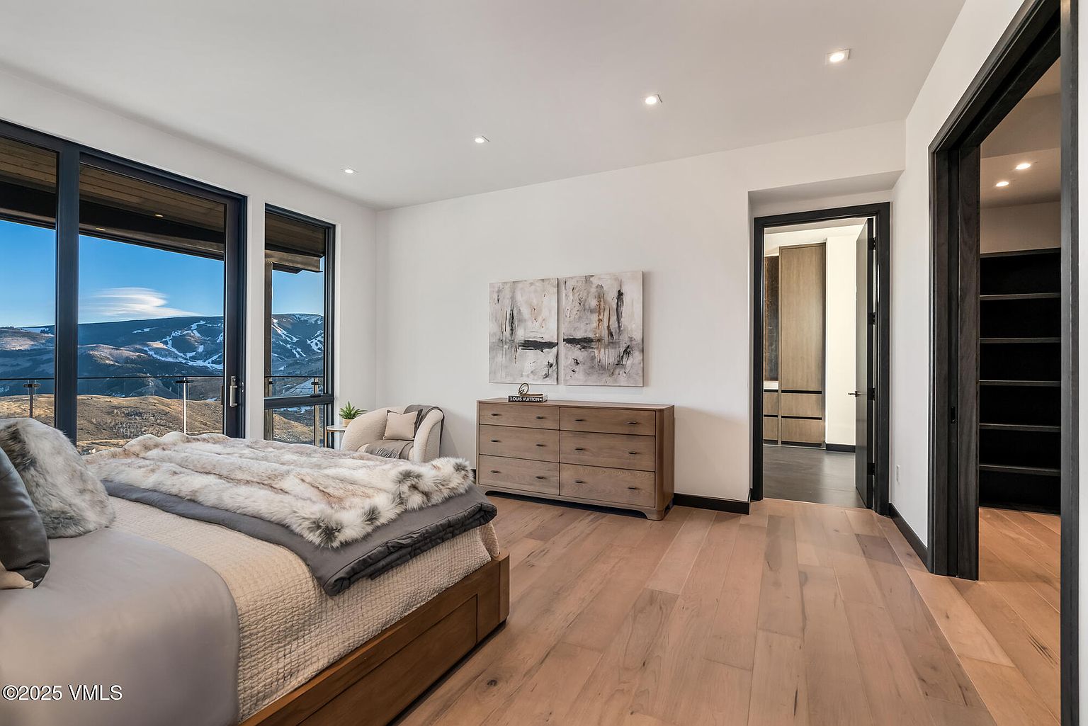 This is a primary bedroom featuring a large bed with a fur throw, offering a luxurious and comfortable feel. The room has large windows with mountain views, a light wood dresser with artwork above, and access to a walk-in closet and another room, suggesting ample storage and space. The hardwood floors and neutral color palette create a serene and inviting atmosphere.