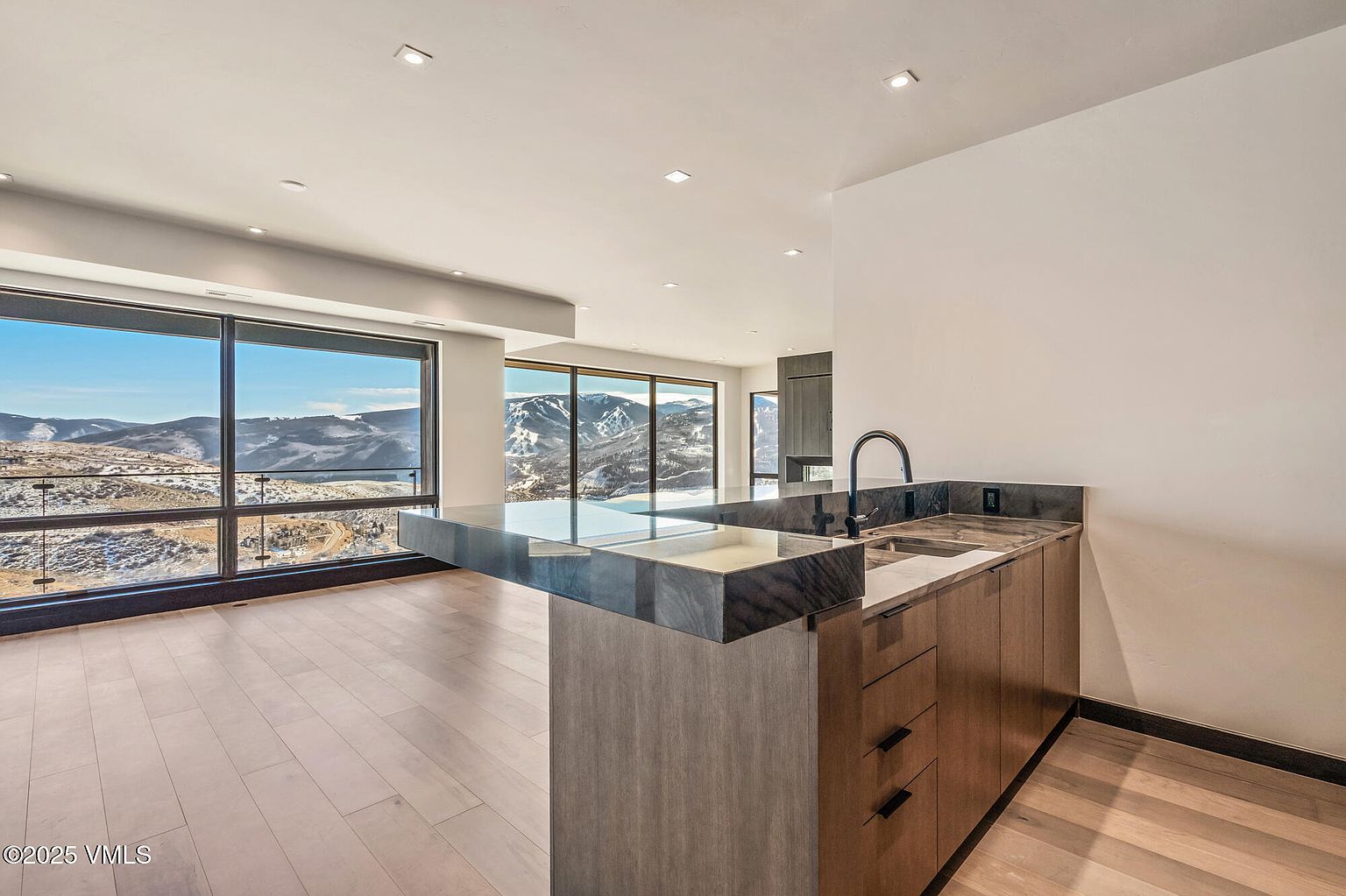 This interior shot showcases a modern kitchen area with a sleek countertop and cabinetry. Large windows offer a stunning view of the snow-covered mountains, bringing natural light into the space. The hardwood flooring and minimalist design contribute to a clean and contemporary aesthetic.