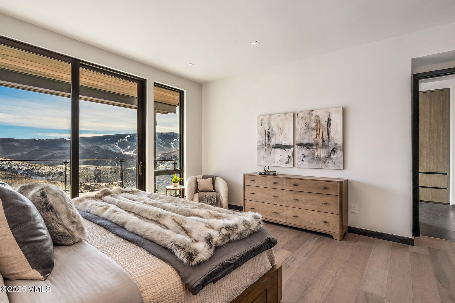 This is a primary bedroom featuring a large bed with a faux fur throw, offering a cozy and luxurious feel. The room has large windows with mountain views, a wooden dresser with artwork above, and hardwood floors. The color palette is neutral, creating a serene and inviting atmosphere.