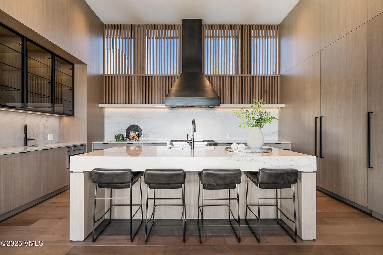 This is a modern kitchen featuring a large island with seating, light wood cabinetry, and a marble backsplash. A striking black range hood is centered above the cooktop, and natural light filters through the slatted window design. The overall impression is one of sophisticated elegance and functionality.