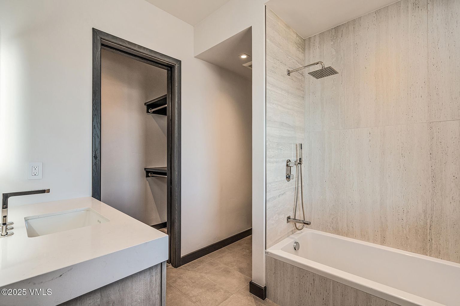 This is a well-lit primary bathroom featuring a modern design. The bathroom includes a vanity with a white countertop and a rectangular sink, a walk-in closet, and a bathtub with a shower. The walls are covered in light-colored tiles, and the overall impression is clean and luxurious.