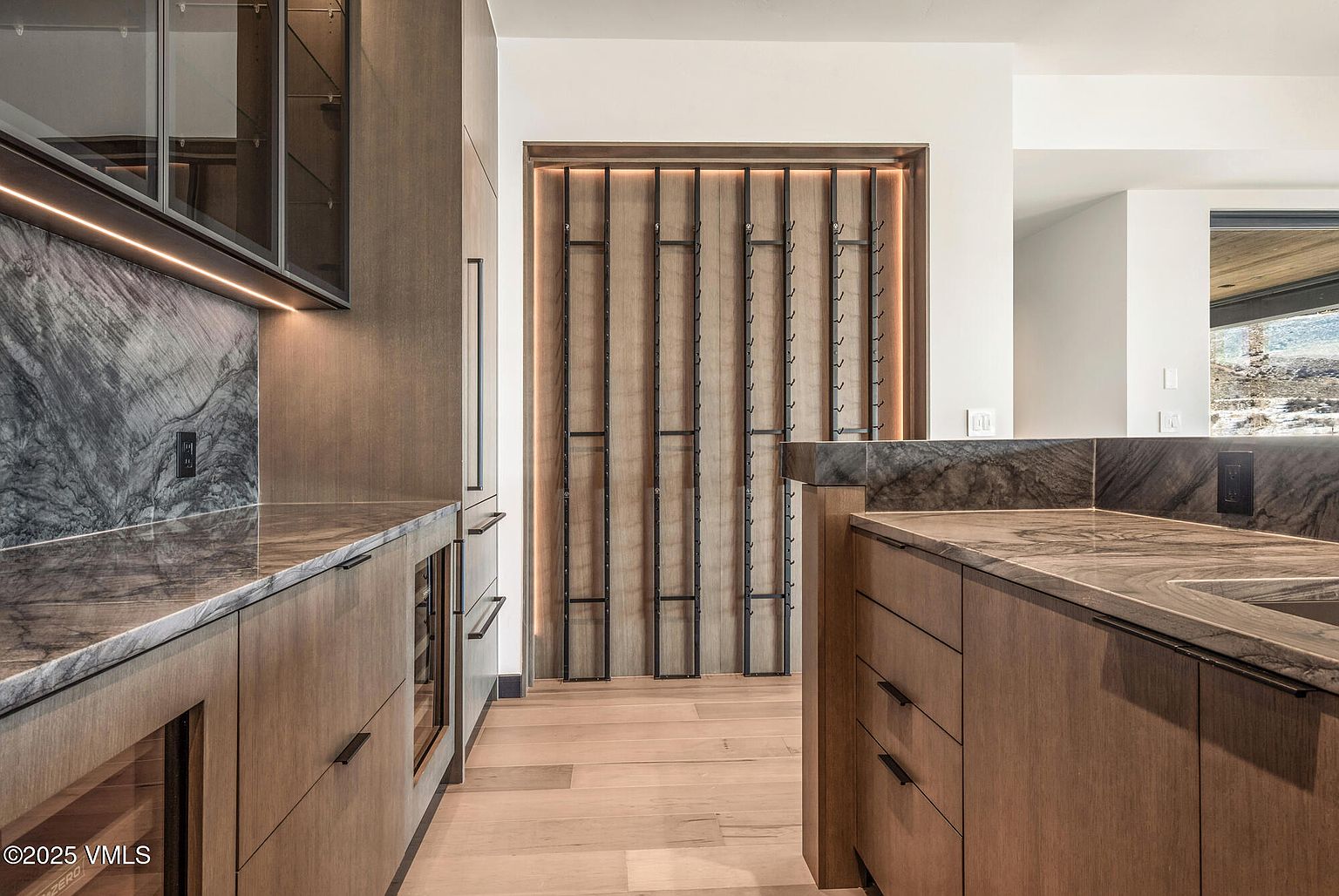 This interior shot showcases a modern kitchen with sleek, wooden cabinetry and marble countertops. A unique wine rack feature is integrated into the wall, adding a touch of sophistication. The hardwood flooring and recessed lighting contribute to the warm and inviting atmosphere of the space.