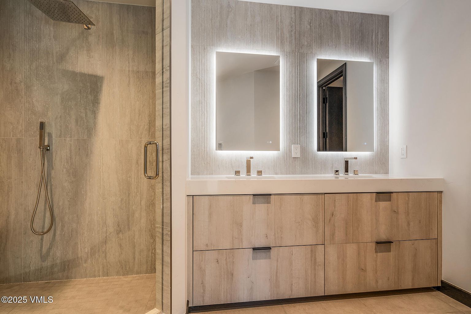 This is a primary bathroom featuring a double vanity with light wood cabinetry and a white countertop. Two illuminated mirrors hang above the sinks, set against a textured gray wall. A glass-enclosed shower is visible to the left, showcasing modern fixtures and tile work, creating a spa-like atmosphere.