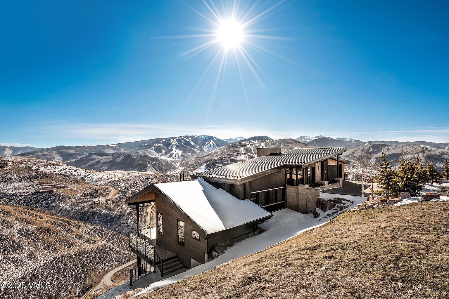 This image showcases a modern mountain home with a striking architectural design. The house features a combination of wood and metal elements, with a partially snow-covered roof. It is situated on a hillside, offering panoramic views of the surrounding snow-dusted mountains under a clear blue sky with a bright sun.