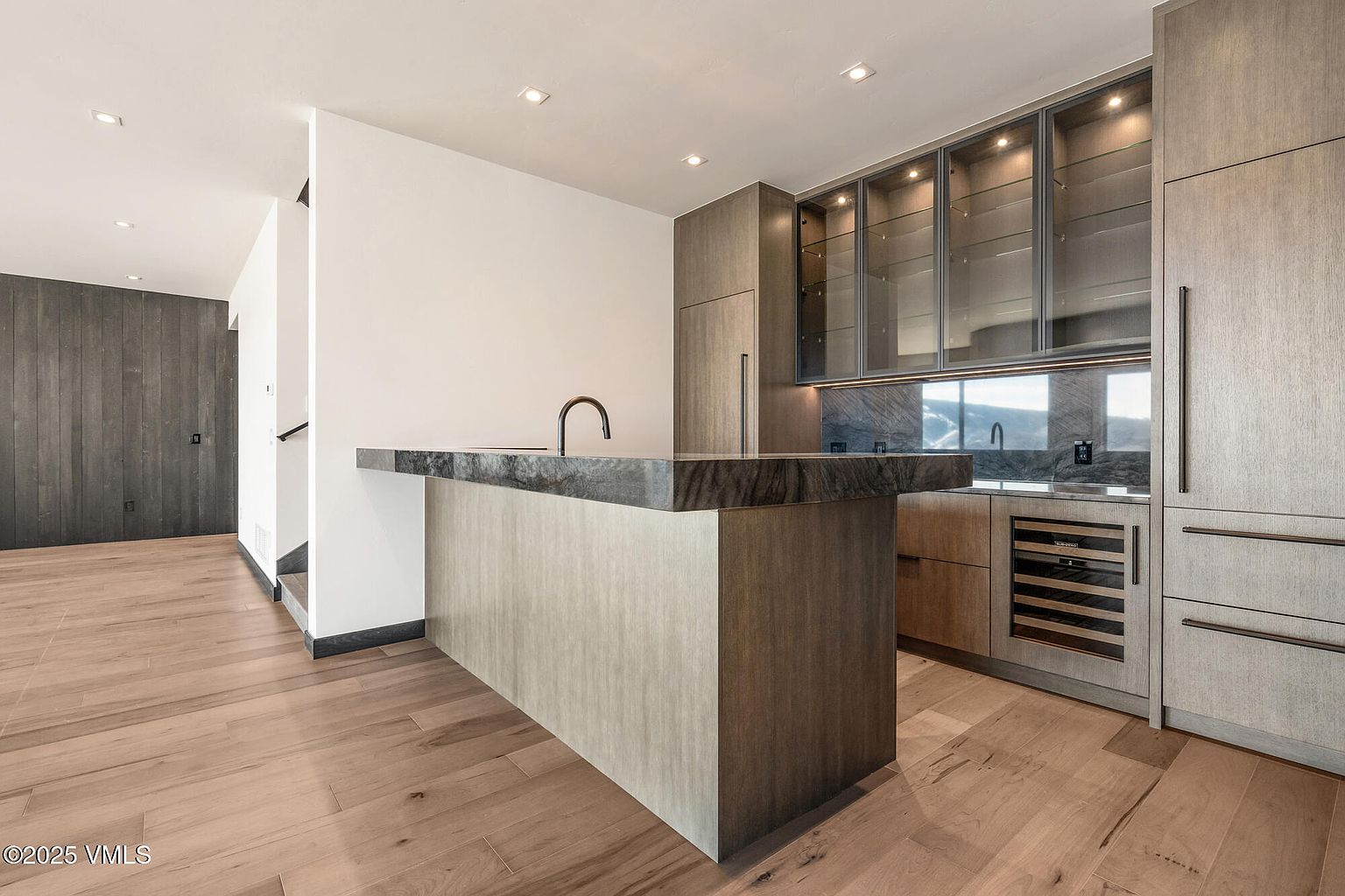 This interior shot showcases a modern kitchen with light wood flooring and cabinetry. A dark countertop island with a sink is prominently featured, complemented by glass-front cabinets with interior lighting. The kitchen also includes a wine cooler and a built-in refrigerator, creating a sleek and functional space.