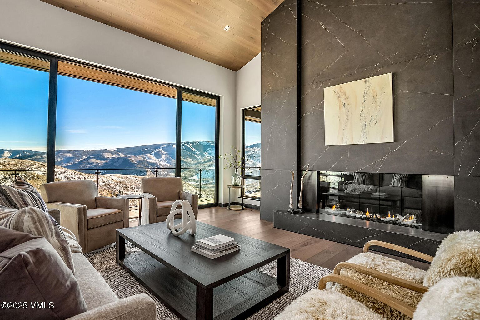 This is a luxurious living room featuring a modern fireplace with a dark marble surround and a contemporary art piece above. Large windows offer stunning views of the mountains, enhancing the room's appeal. The space is furnished with comfortable seating and a stylish coffee table, creating an inviting and sophisticated atmosphere.