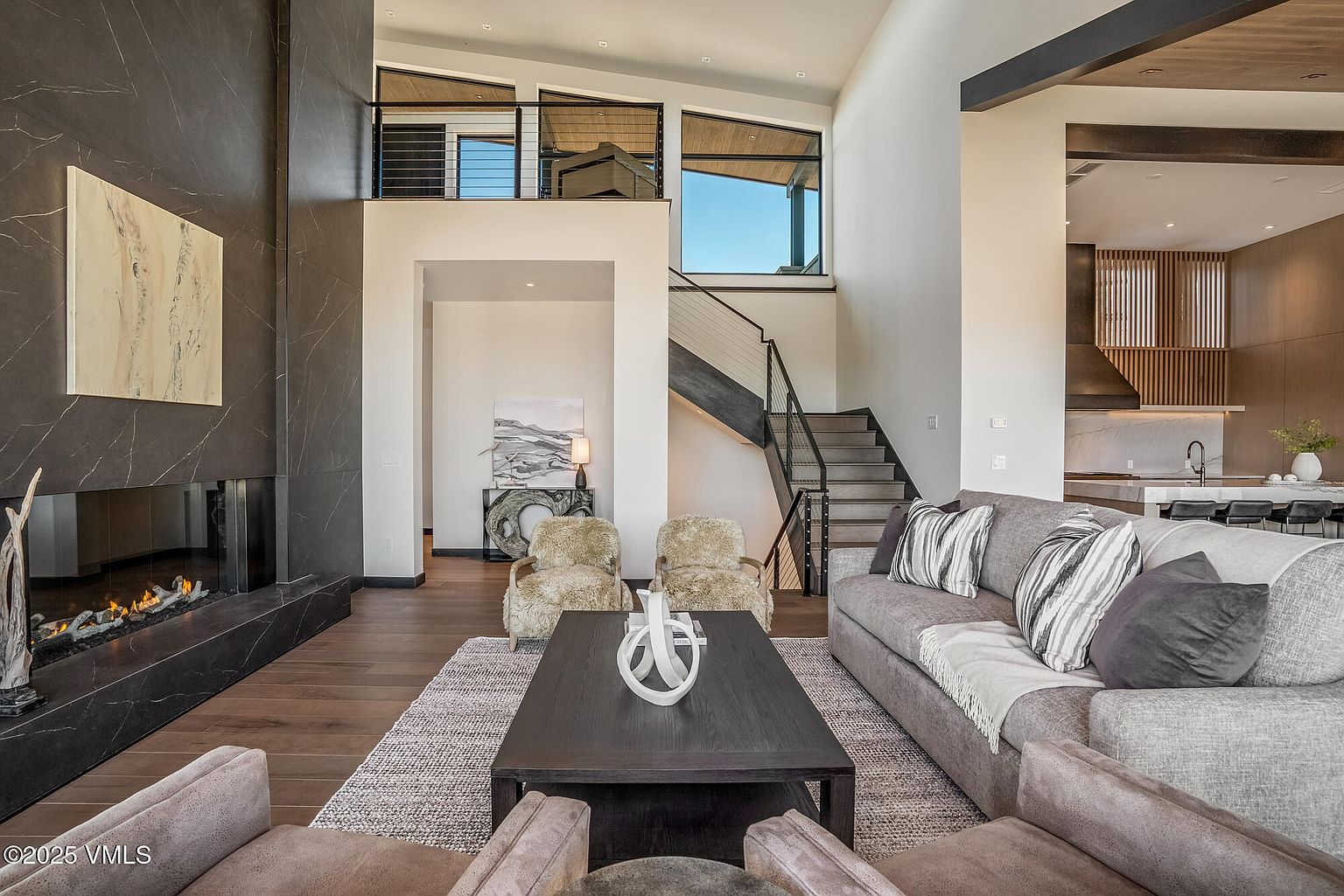 This is a spacious and modern living room featuring a dark marble fireplace, hardwood floors, and a neutral color palette. The room is well-lit with natural light and includes comfortable seating arrangements with a large sofa and armchairs. A staircase leads to an upper level, adding architectural interest and highlighting the home's open design.