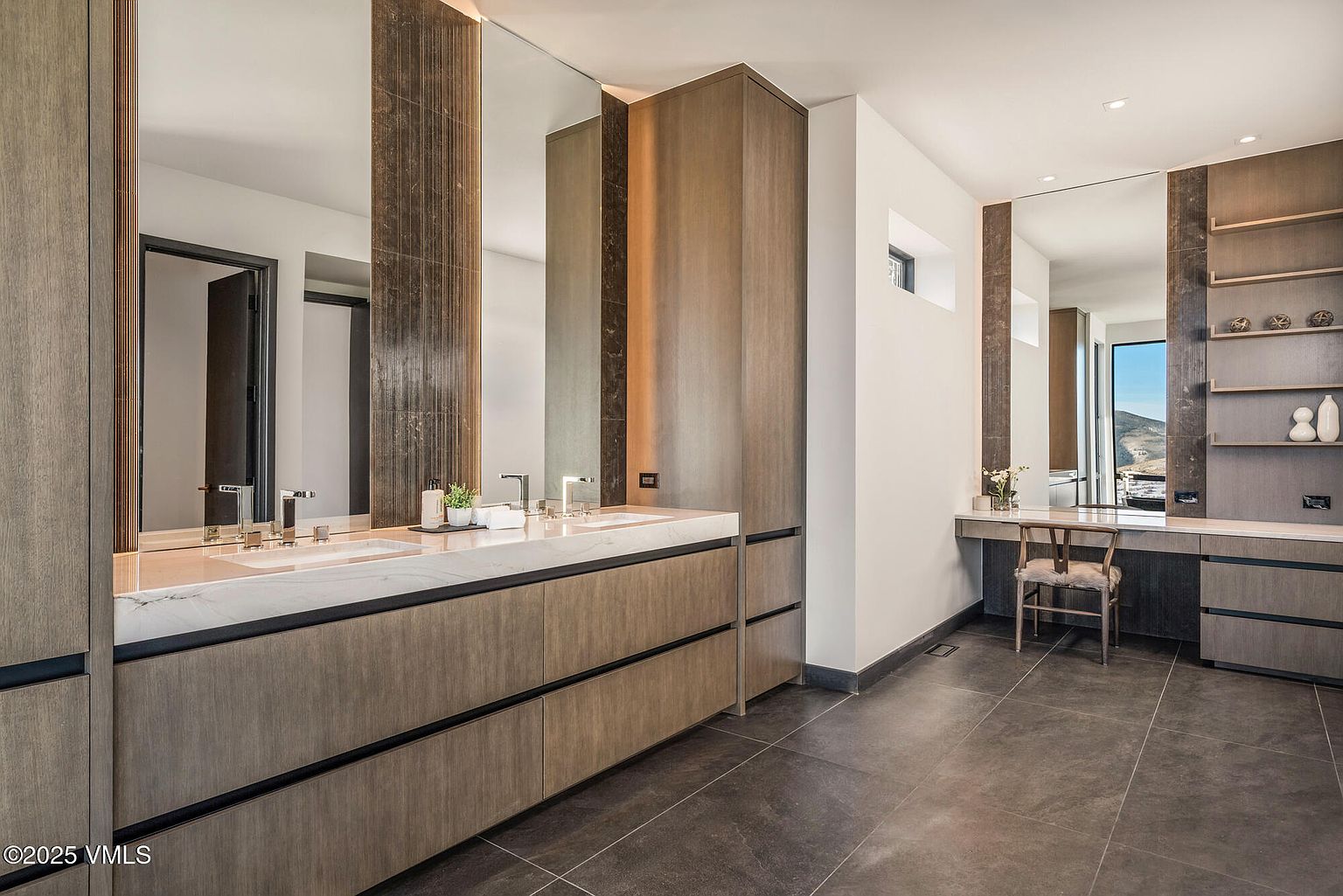 This is a luxurious primary bathroom featuring a double vanity with a marble countertop and modern faucets. The cabinetry is a warm wood tone with sleek, dark accents. A separate vanity area with a chair and shelving provides a dedicated space for grooming, and the dark tile flooring adds a touch of sophistication.