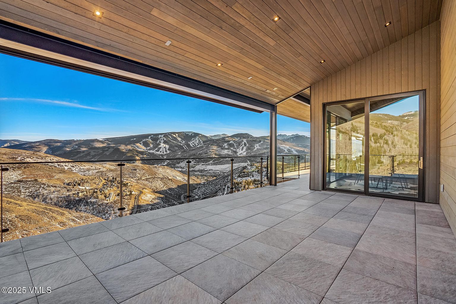 This image showcases a luxurious outdoor patio or balcony with stunning mountain views. The space features a modern design with sleek glass railings, large format tile flooring, and a seamless transition to the interior through a sliding glass door. The wooden ceiling adds warmth and sophistication to the outdoor living area.