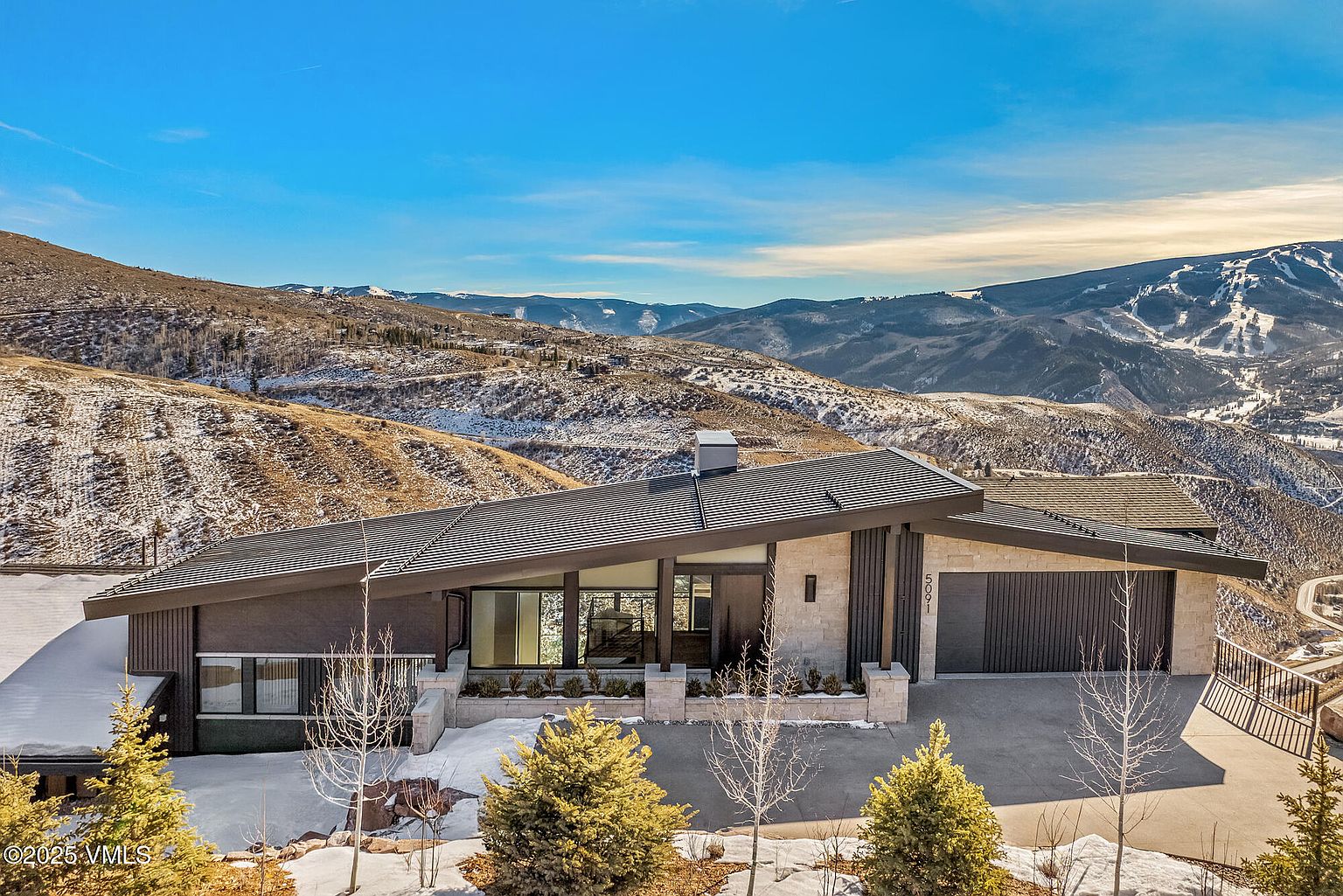 This is a front exterior view of a modern home nestled in a mountainous landscape. The house features a unique roofline, a combination of stone and dark wood siding, and large windows that offer stunning views. The property is surrounded by natural terrain with sparse vegetation and snow patches, creating a serene and private setting.