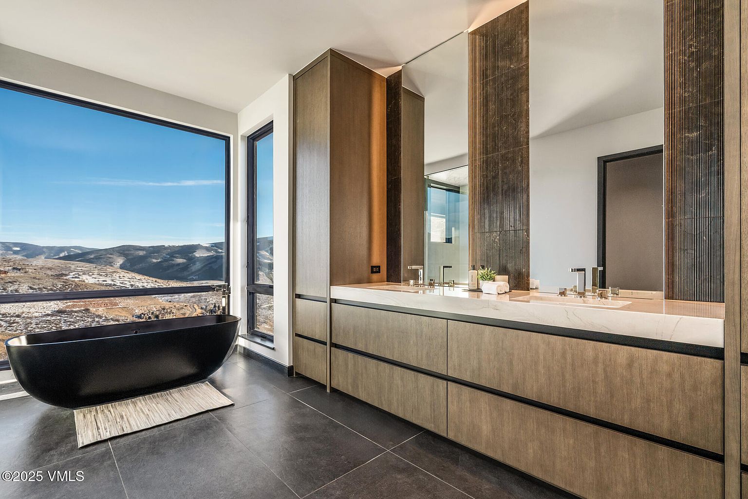 This is a luxurious primary bathroom featuring a modern freestanding black bathtub positioned in front of a large window with mountain views. The bathroom includes a double vanity with sleek wood cabinetry, a marble countertop, and contemporary fixtures. The overall design is minimalist and elegant, emphasizing natural light and high-end finishes.
