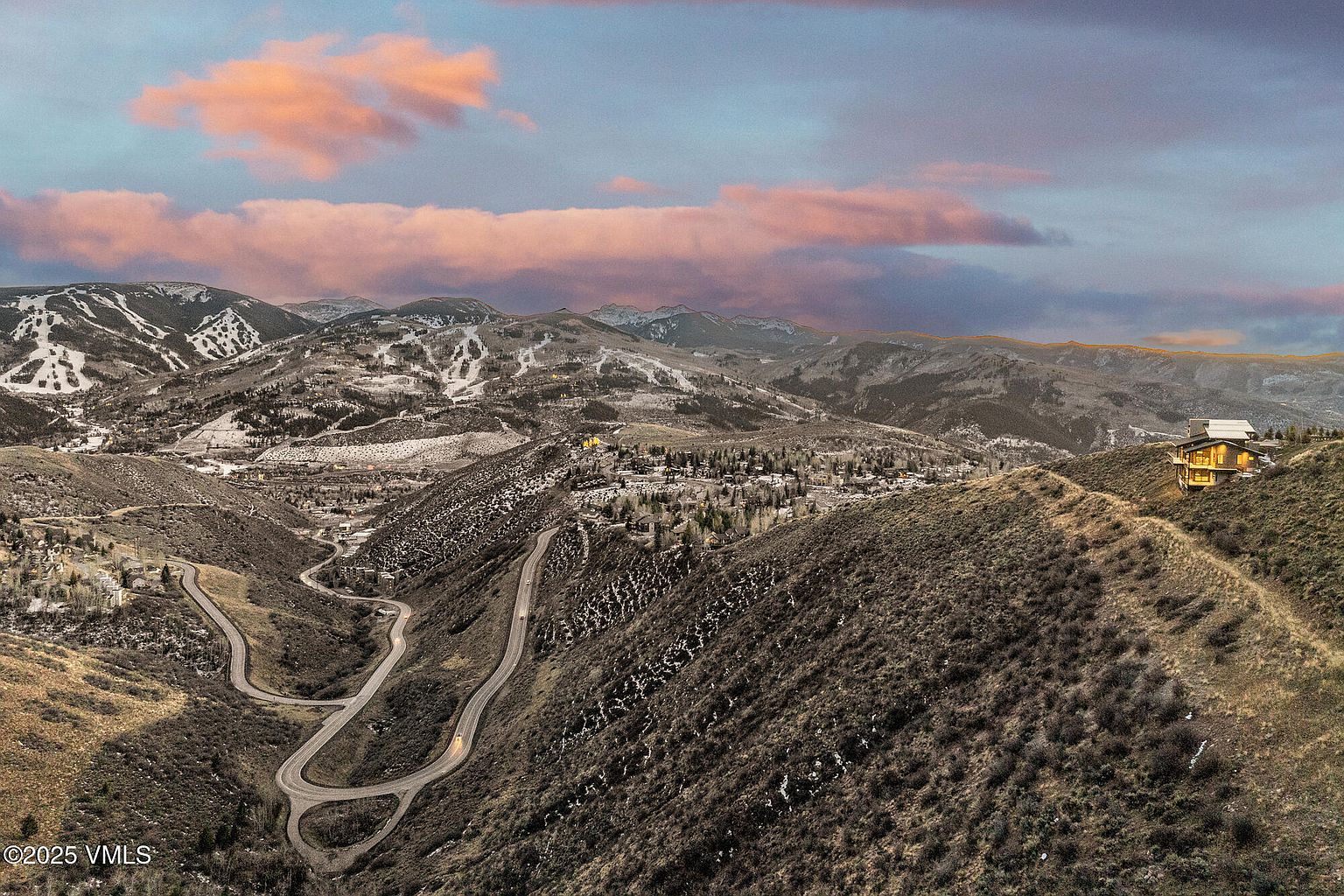 This aerial view showcases a luxurious home perched atop a hill, accessible via a winding road. The surrounding landscape features rolling hills with sparse vegetation and snow-capped mountains in the distance, creating a sense of seclusion and natural beauty. The home's architecture blends seamlessly with the environment, offering a unique and desirable property.