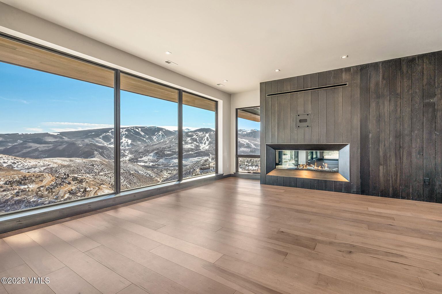 This is an interior shot of a modern living room featuring a large window with a scenic mountain view. The room has hardwood floors and a contemporary fireplace with dark wood paneling. The space is bright and airy, emphasizing the connection to the outdoors.