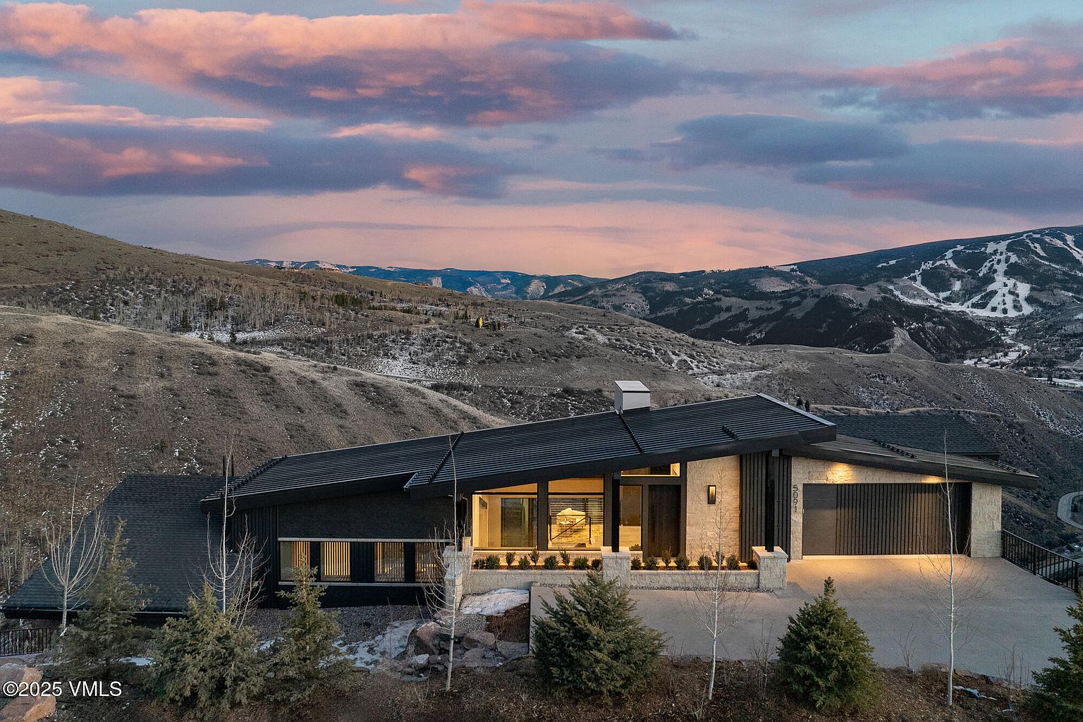 This is an exterior front view of a modern home nestled in a mountainous landscape. The house features a dark roof, a mix of dark siding and stone accents, and large windows that offer ample natural light. The property is surrounded by natural terrain, with mountains visible in the background under a dramatic sky, suggesting a serene and private setting.