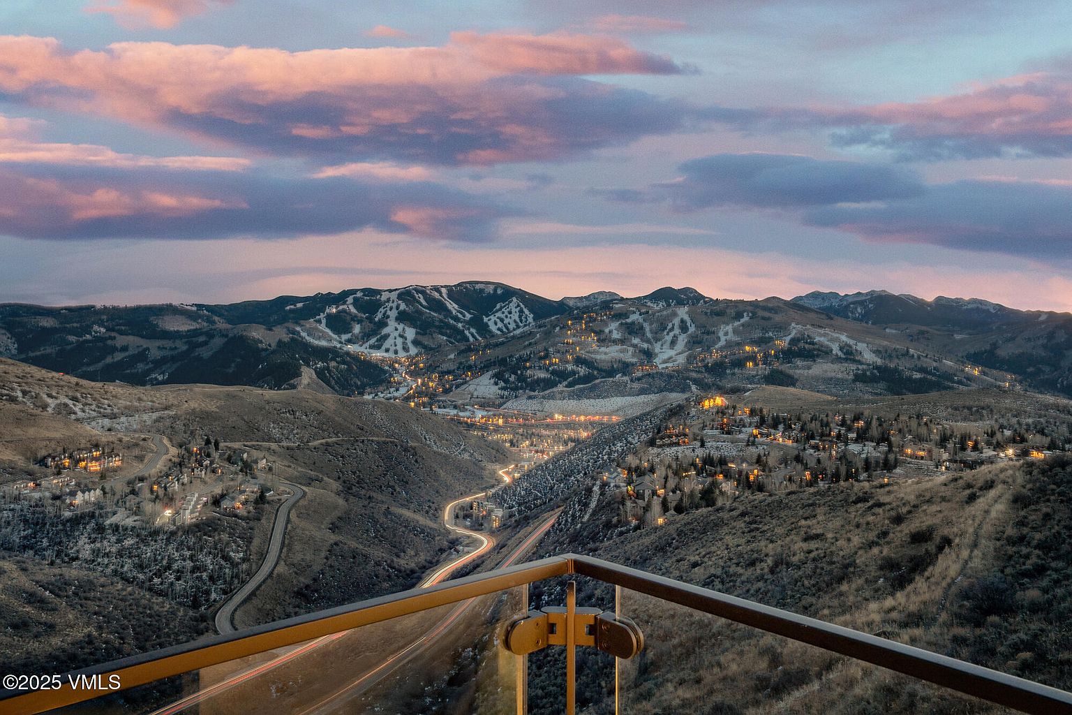 This aerial view showcases a stunning mountain landscape at dusk, with a winding road leading through the valley and illuminated houses dotting the hillsides. The sky is painted with hues of pink and purple, creating a serene and picturesque scene. A balcony railing in the foreground offers a sense of perspective and highlights the elevated vantage point.