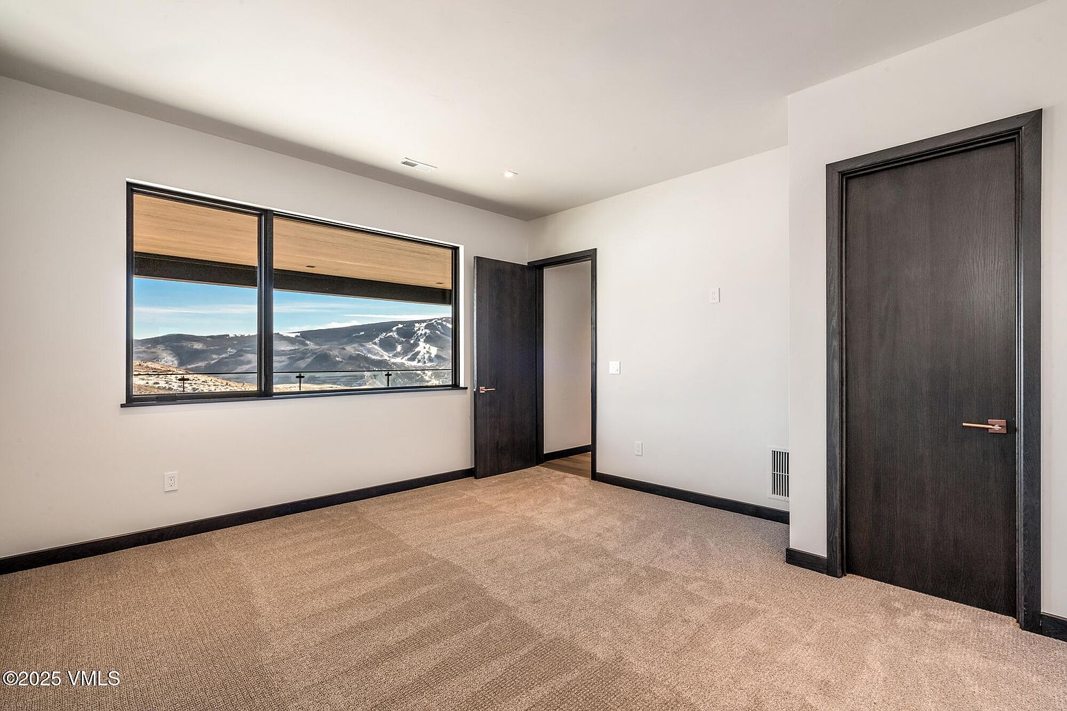 This is a well-lit bedroom featuring neutral-toned carpet and walls. A large window offers a scenic view of snow-capped mountains. The room includes two dark-stained doors with matching trim, adding a touch of modern elegance.