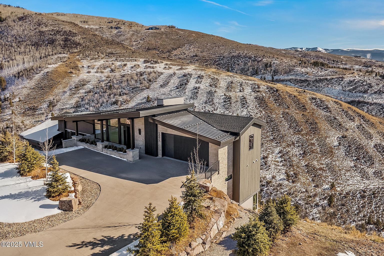 This image showcases the front exterior of a modern home nestled on a hillside. The architectural design features a combination of stone and wood siding, with a sleek, angled roofline. A concrete driveway leads up to the garage, and the surrounding landscape includes mature trees and natural terrain, creating a sense of privacy and integration with the environment.