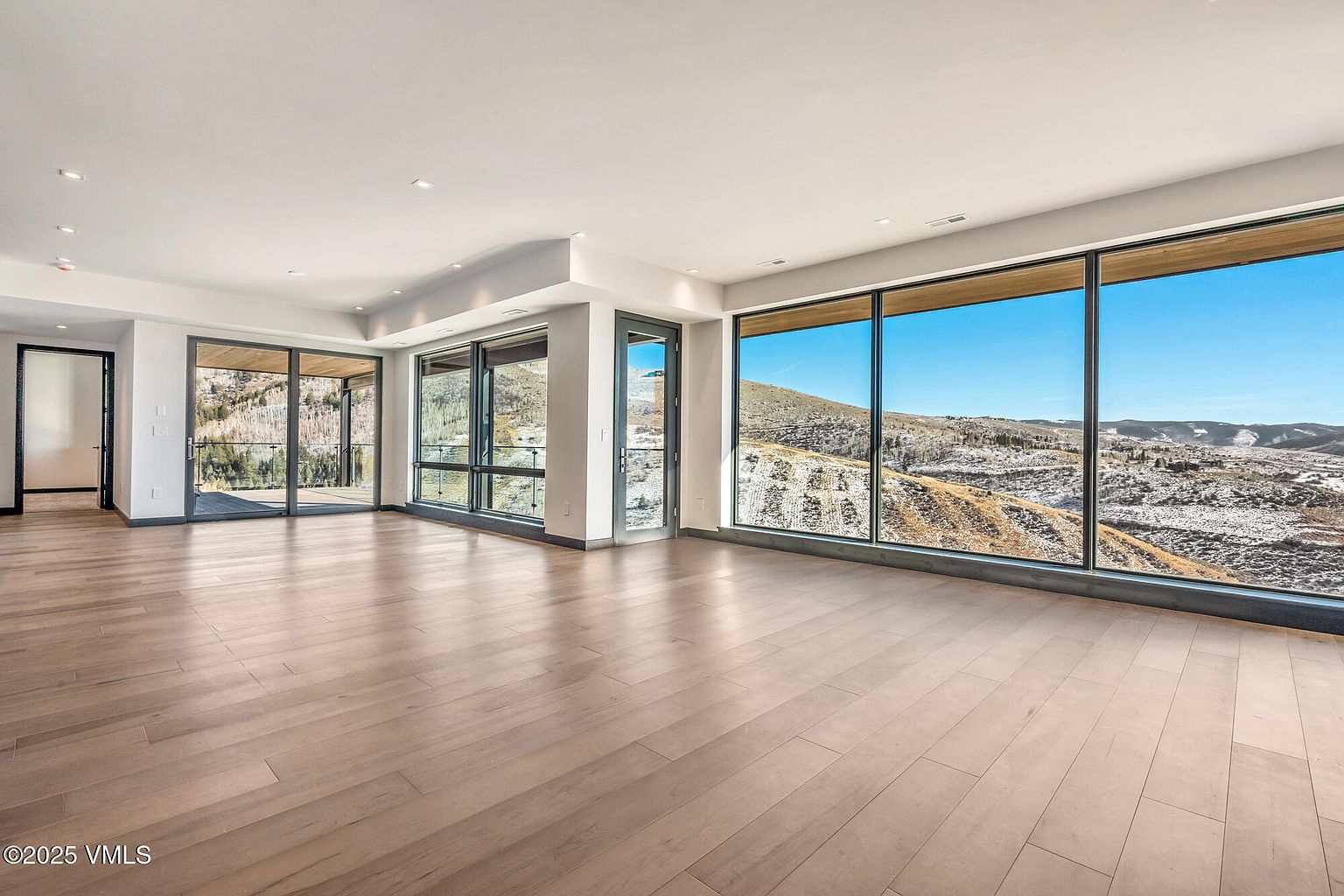 This is a spacious living room featuring light wood flooring and large windows that offer a panoramic view of a mountainous landscape. The room is bright and airy, with a minimalist design that emphasizes the natural light and scenic views. The open floor plan and neutral color palette create a sense of calm and sophistication.