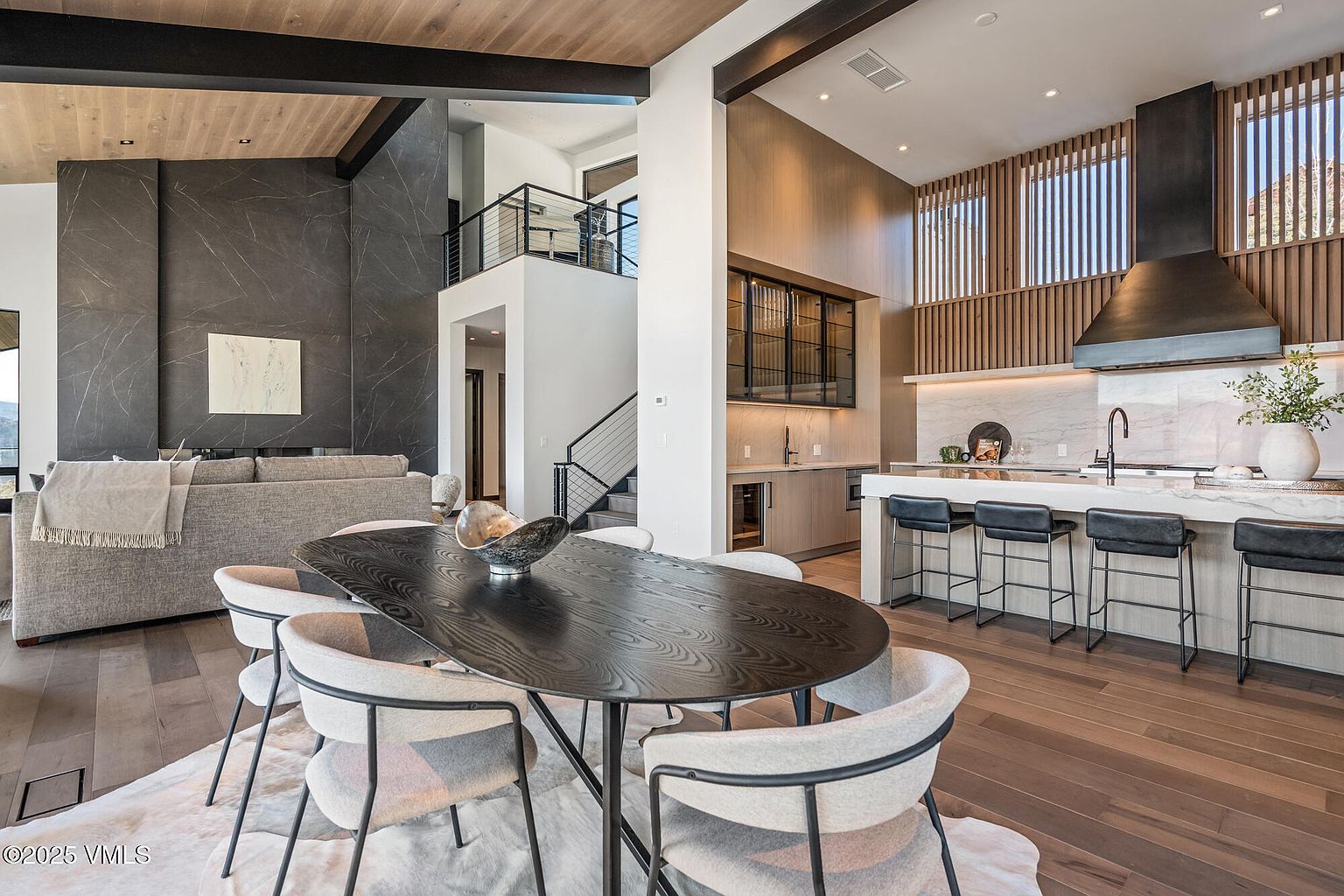 This interior shot showcases a modern dining area adjacent to a kitchen and living space. The dining area features a dark wood oval table surrounded by light-colored chairs on a white rug. The open-concept design highlights the home's high ceilings, natural wood accents, and contemporary style, creating a luxurious and inviting atmosphere.