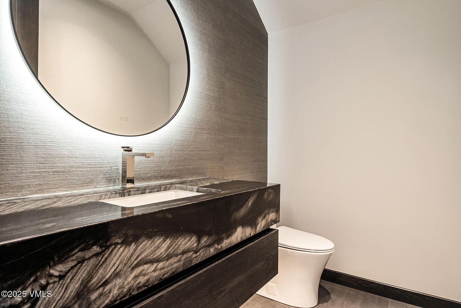 This is a modern guest bathroom featuring a sleek, dark vanity with a marble countertop and an integrated sink. A round, illuminated mirror hangs above the vanity, complementing the textured gray wallpaper. A white toilet is positioned to the right, and the overall design is minimalist and stylish.
