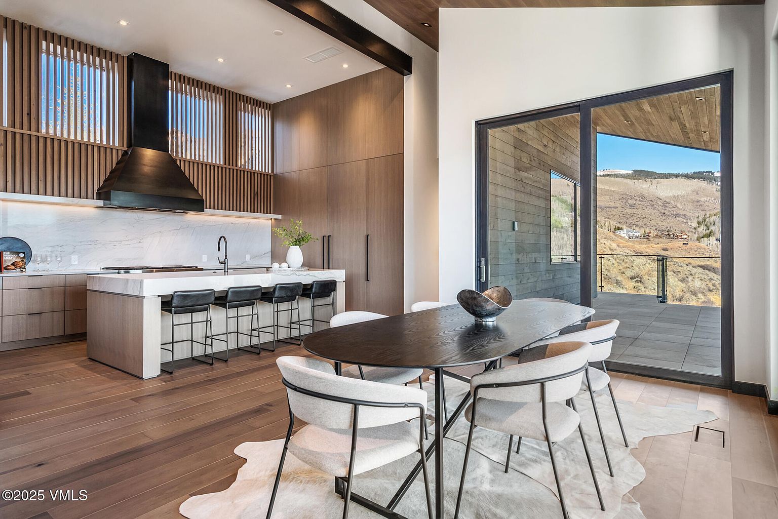 This interior shot showcases a modern dining area adjacent to a kitchen. The dining space features a dark wood oval table surrounded by six chairs with light-colored upholstery and dark metal frames, set upon a cowhide rug. A sliding glass door provides access to an outdoor deck with mountain views, enhancing the room's appeal and natural light.
