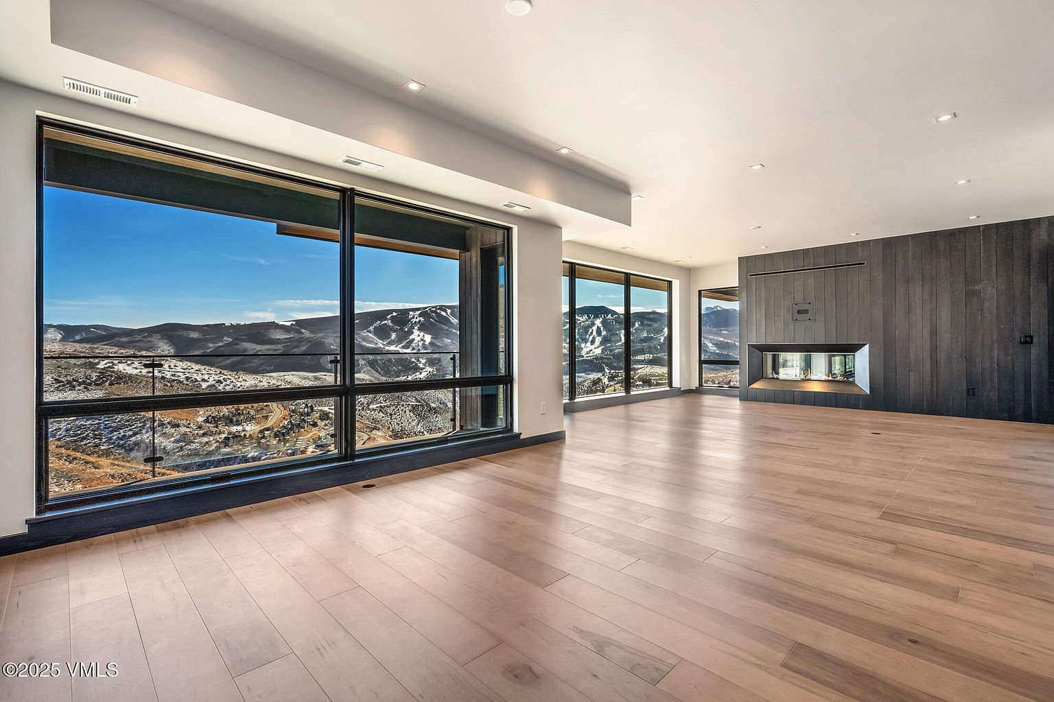 This is a spacious living room featuring large windows with mountain views. The room has hardwood floors and a modern fireplace with a dark wood surround. The overall impression is one of luxury and sophistication, with a focus on natural light and scenic views.