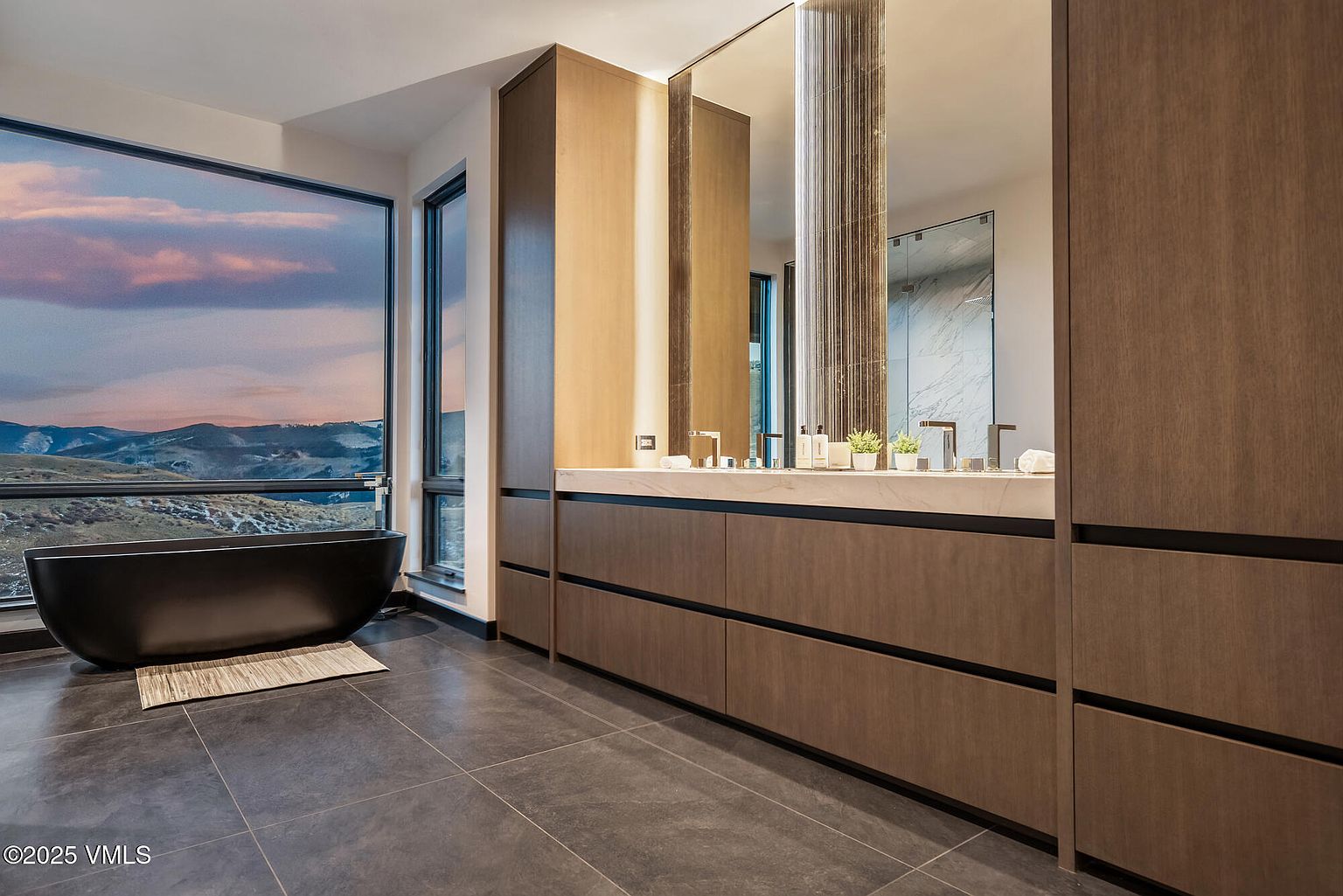 This is a luxurious primary bathroom featuring a modern design with dark wood cabinetry and a double vanity with a light countertop. A freestanding black bathtub sits by a large window offering scenic mountain views. The bathroom has a sleek and sophisticated aesthetic, perfect for relaxation and comfort.