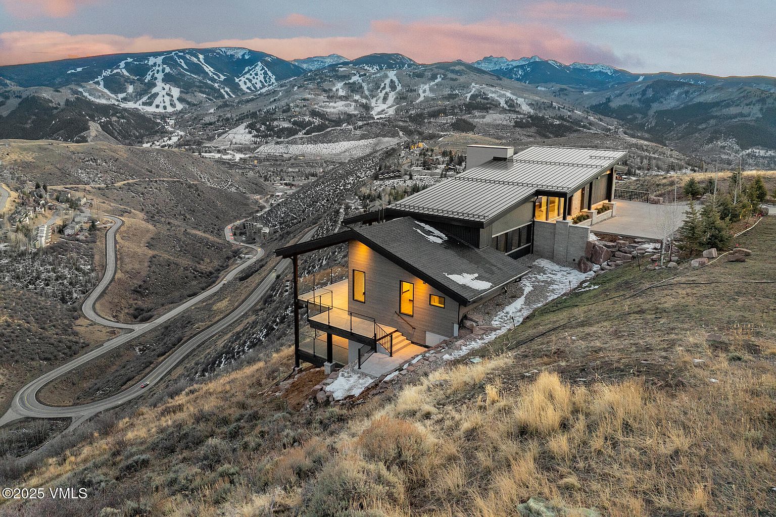 This aerial view showcases a modern home nestled on a hillside, featuring a sleek, multi-level design with a dark roof and expansive windows. A winding road leads up to the property, and the backdrop includes stunning mountain views with ski slopes. The landscape is a mix of natural terrain and manicured areas, highlighting the home's integration with its environment.