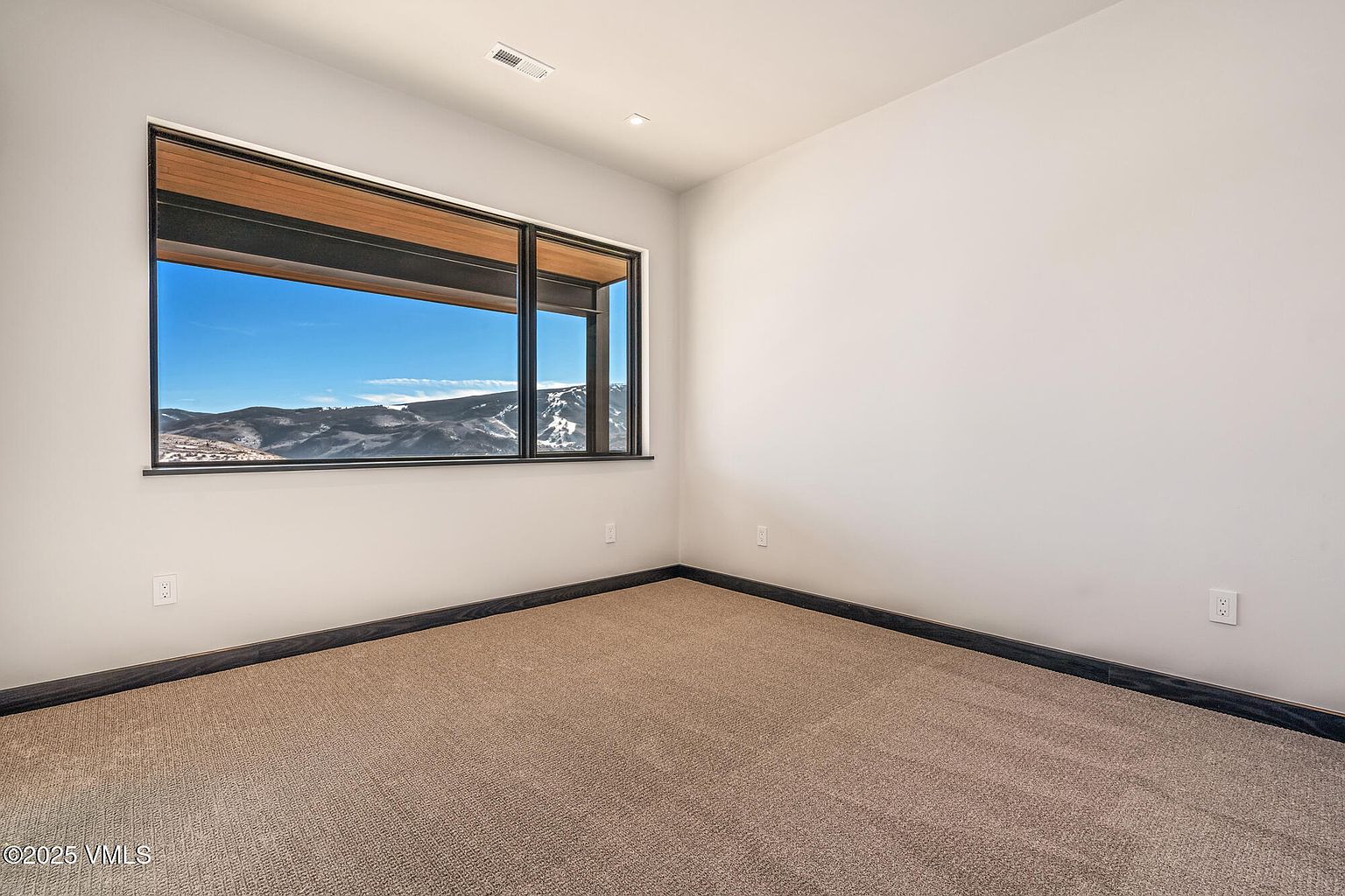 This is an interior shot of a bedroom, likely a guest or secondary bedroom. The room features neutral-toned carpet, light-colored walls, and a large window offering a view of a mountainous landscape. The room is simple and clean, providing a blank canvas for potential buyers to envision their own style.
