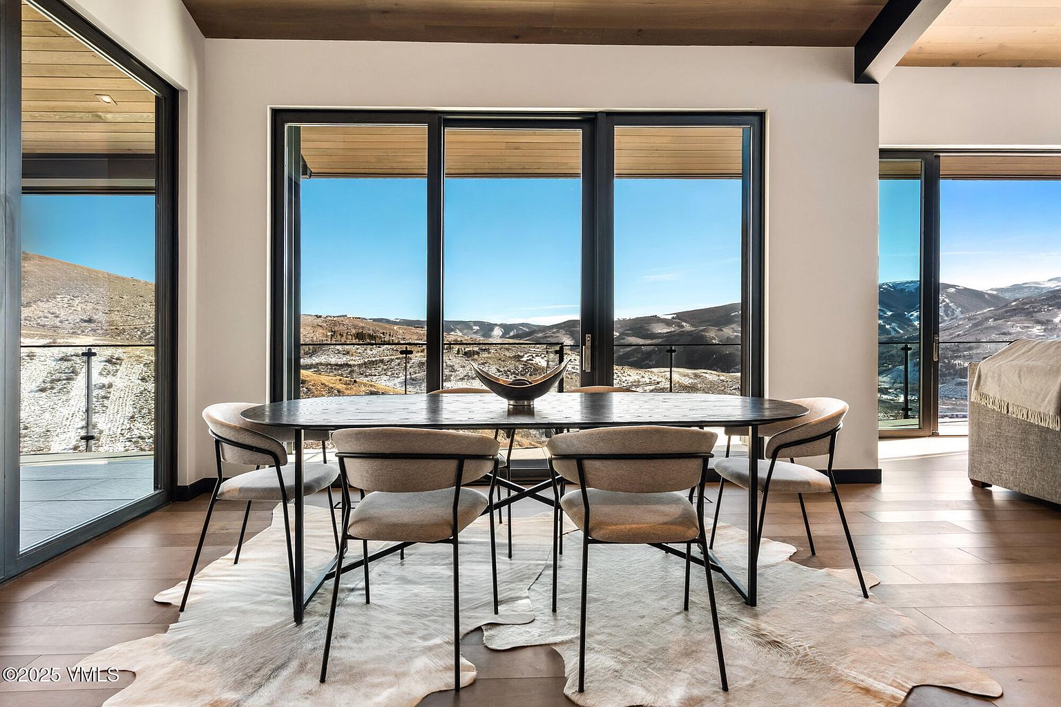 This dining room features a modern aesthetic with a dark oval table surrounded by six chairs with light-colored upholstery and black metal frames. The room is bright, with large windows offering a scenic mountain view, and a faux cowhide rug adds texture to the hardwood floor. The space is well-lit and inviting, perfect for showcasing the home's elegant design.