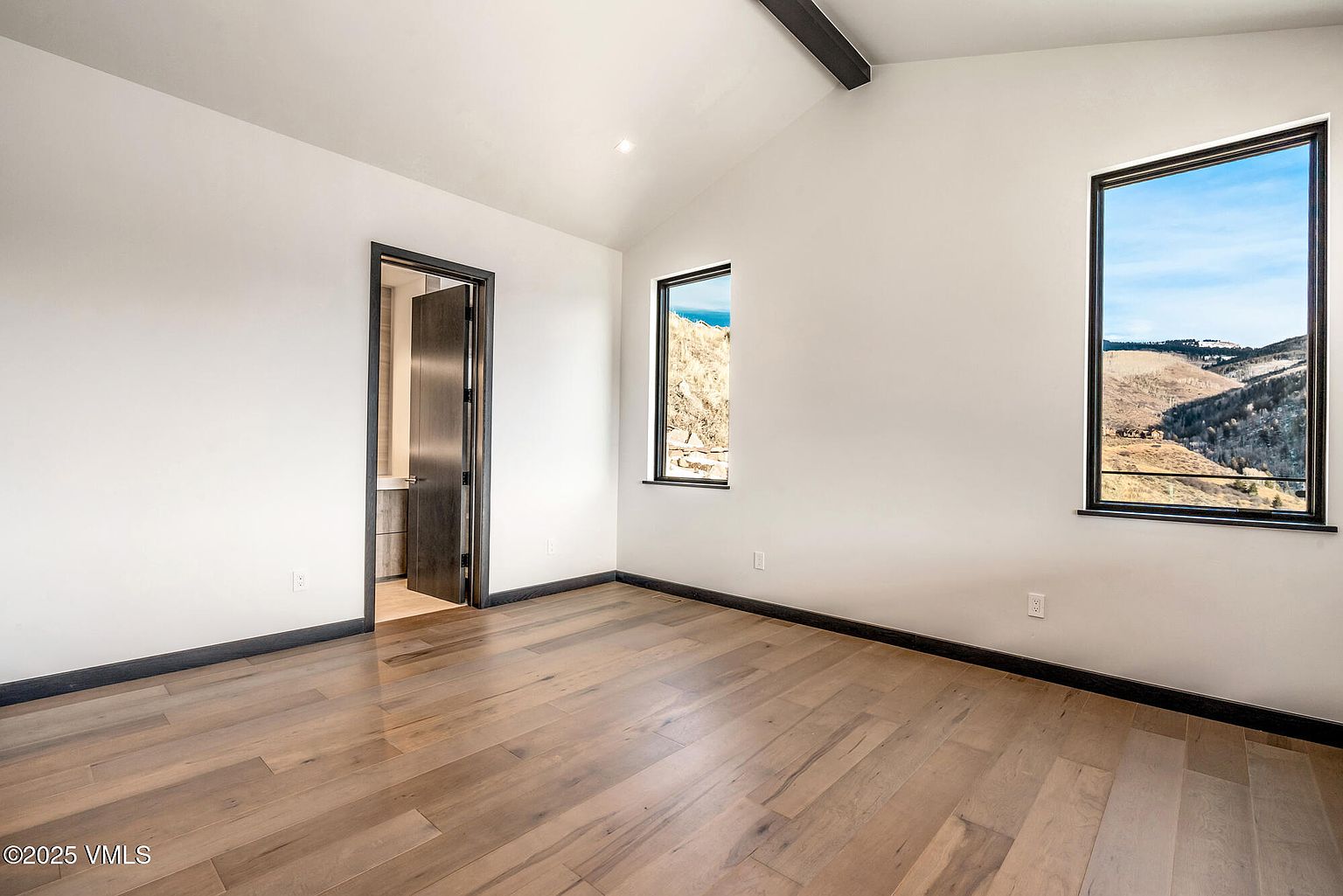 This is an interior shot of a bedroom featuring hardwood floors, white walls, and dark trim. The room has two windows offering natural light and views of the landscape. A doorway leads to an adjacent room, possibly a bathroom or closet, enhancing the sense of space and flow.