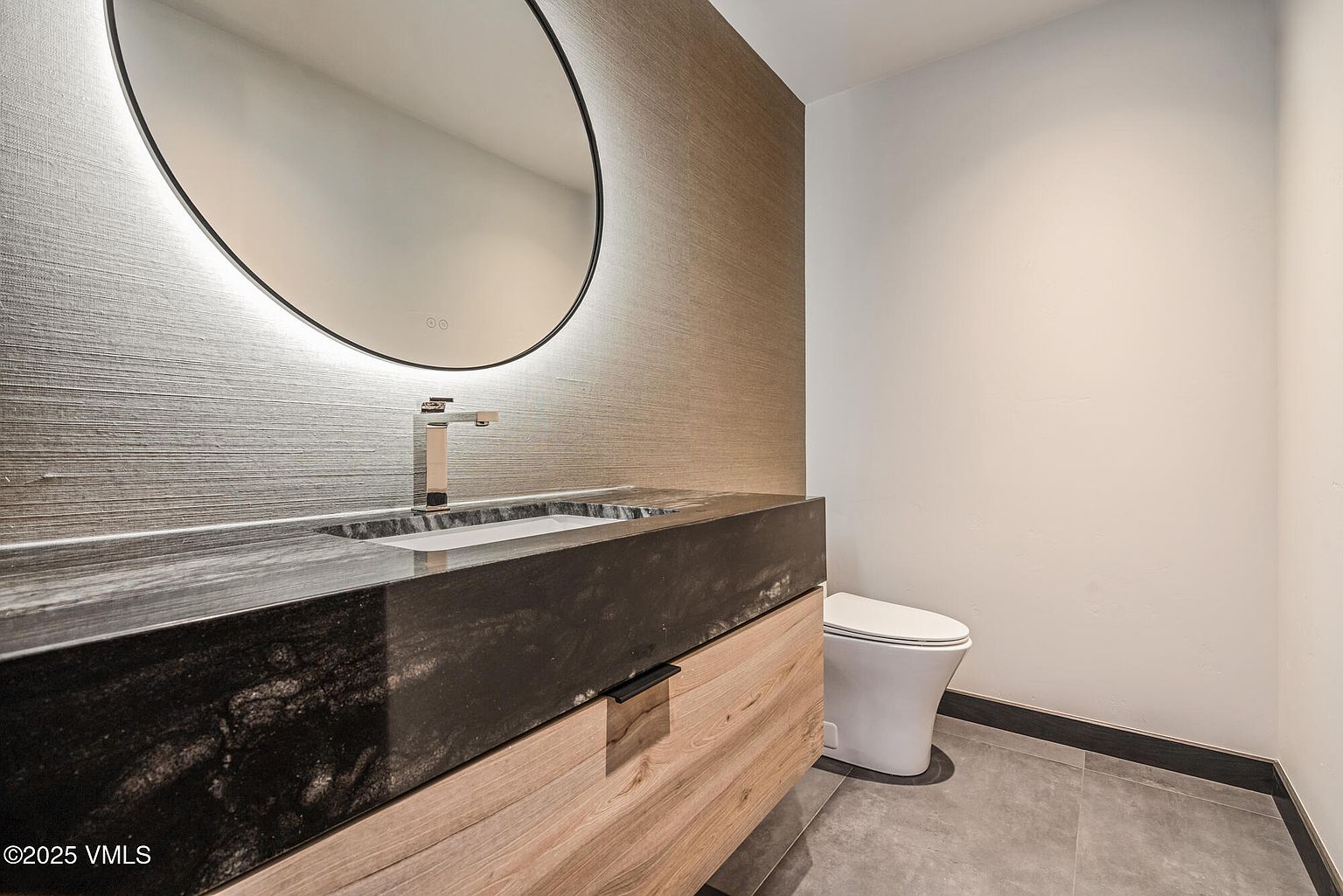 This is a modern guest bathroom featuring a sleek, dark countertop with a built-in sink and a contemporary faucet. A round, illuminated mirror hangs above the sink, complementing the textured wallpaper. A white toilet is visible to the right, and the floor is covered in gray tiles, creating a clean and stylish space.