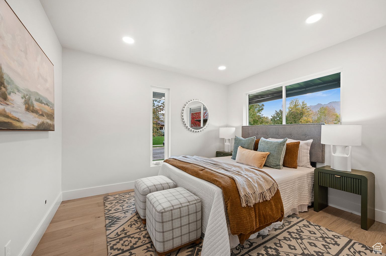 This is a bright and airy primary bedroom featuring a neutral color palette with pops of color from the pillows and throw blanket. The room includes a large window providing natural light, a comfortable bed with a padded headboard, and matching nightstands with lamps. The overall impression is clean, modern, and inviting.