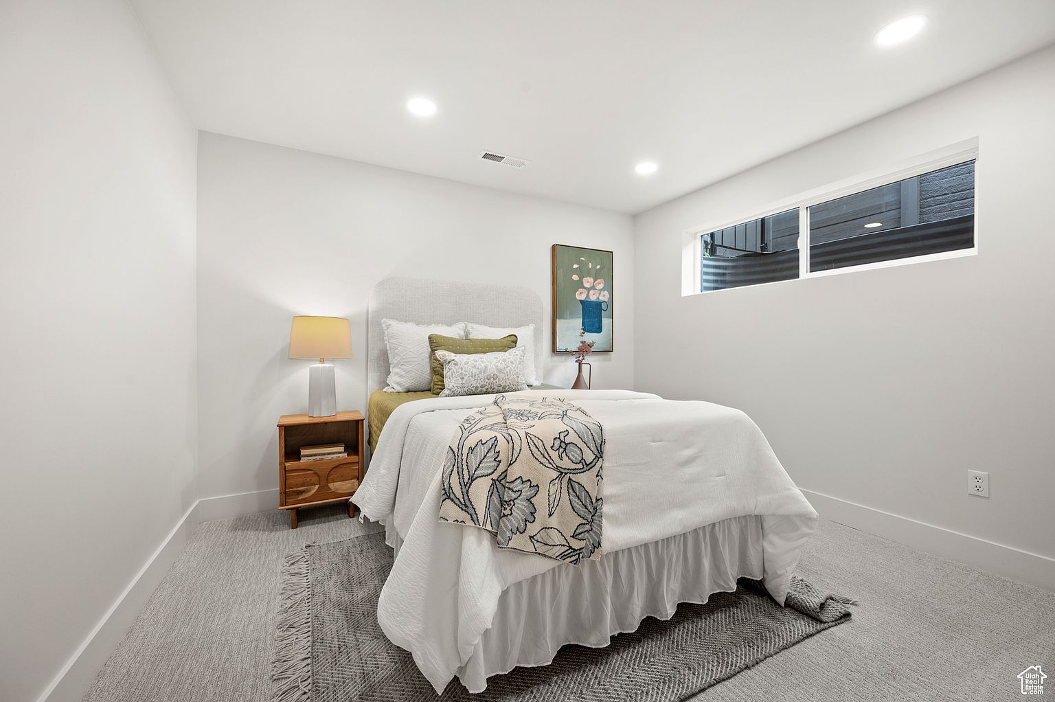This is a cozy bedroom featuring a bed with a light-colored headboard and white bedding accented with a floral throw. A wooden nightstand with a lamp sits beside the bed, and a framed artwork hangs on the wall. The room is painted in a neutral tone, creating a serene and inviting atmosphere, complemented by the soft gray carpet.