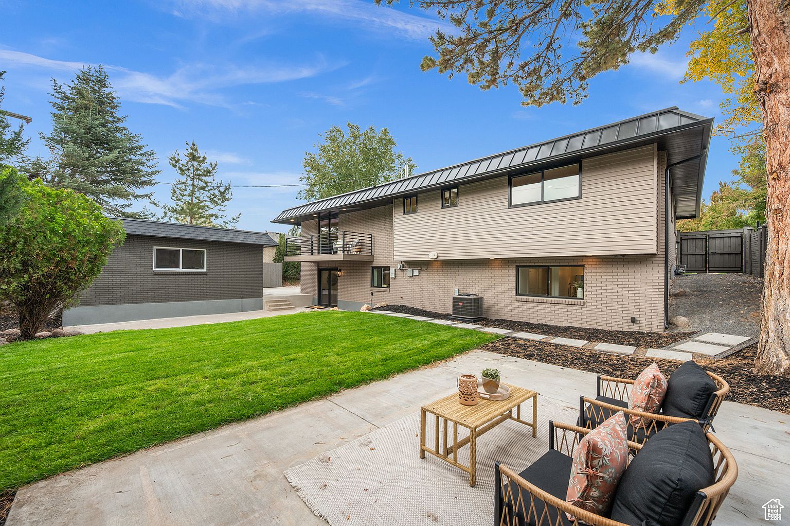 This is a rear view of a modern two-story home with a black metal roof and a combination of brick and siding exterior. The backyard features a well-maintained lawn, a concrete patio with outdoor seating, and a detached garage. The landscaping includes mature trees and shrubs, enhancing the property's appeal.