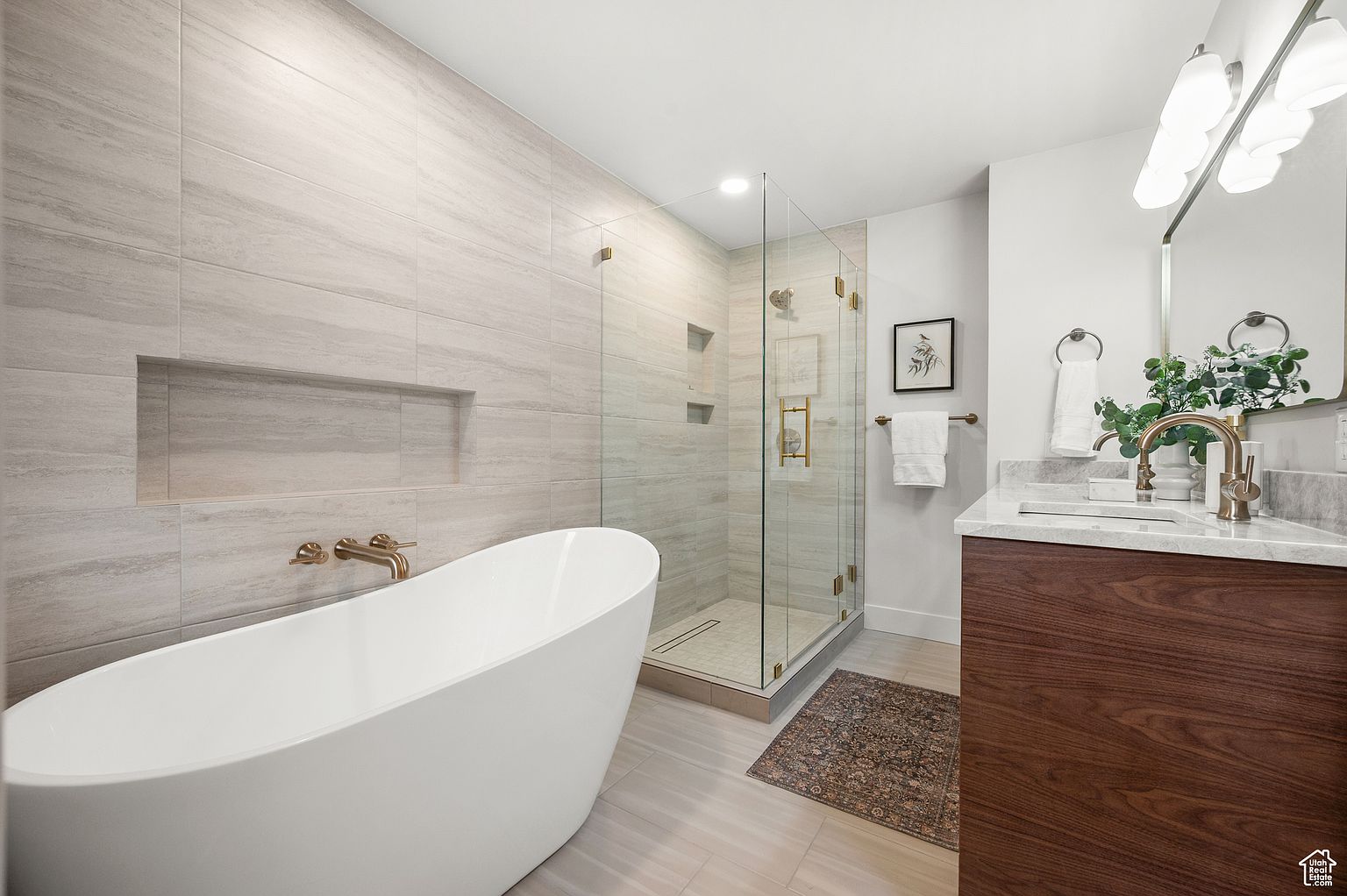 This is a luxurious primary bathroom featuring a freestanding white bathtub with gold fixtures, a glass-enclosed shower with matching gold hardware, and a modern vanity with a marble countertop and dark wood cabinetry. The walls are tiled in a light, neutral tone, and a decorative rug adds warmth to the space. The overall impression is one of elegance and relaxation.