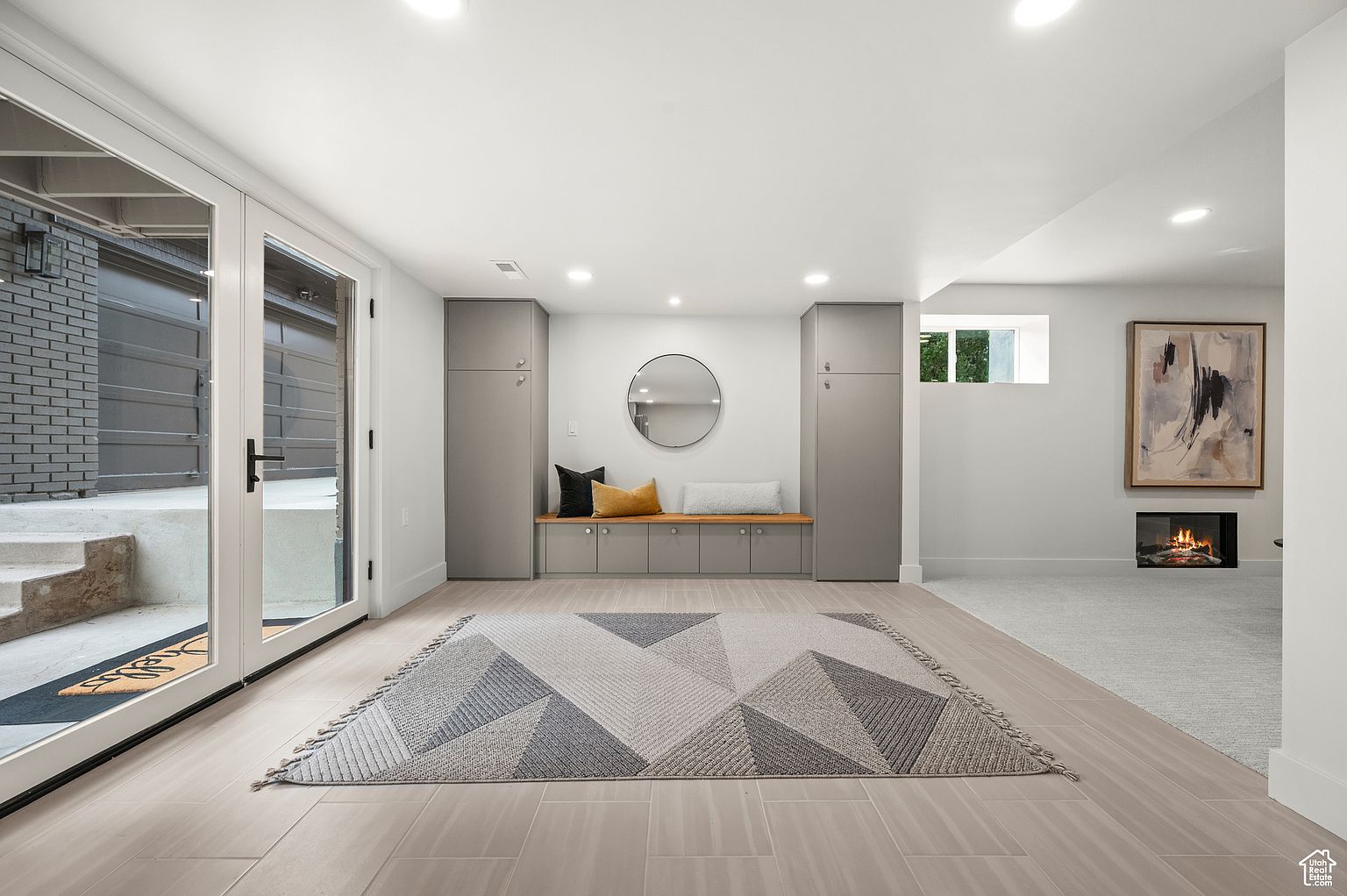 This is a well-lit basement area featuring light-colored tile flooring and a geometric patterned rug. Built-in gray cabinets and a bench with decorative pillows provide storage and seating. A modern fireplace and artwork add a touch of elegance to the space, which is accessible through glass doors leading to the exterior.