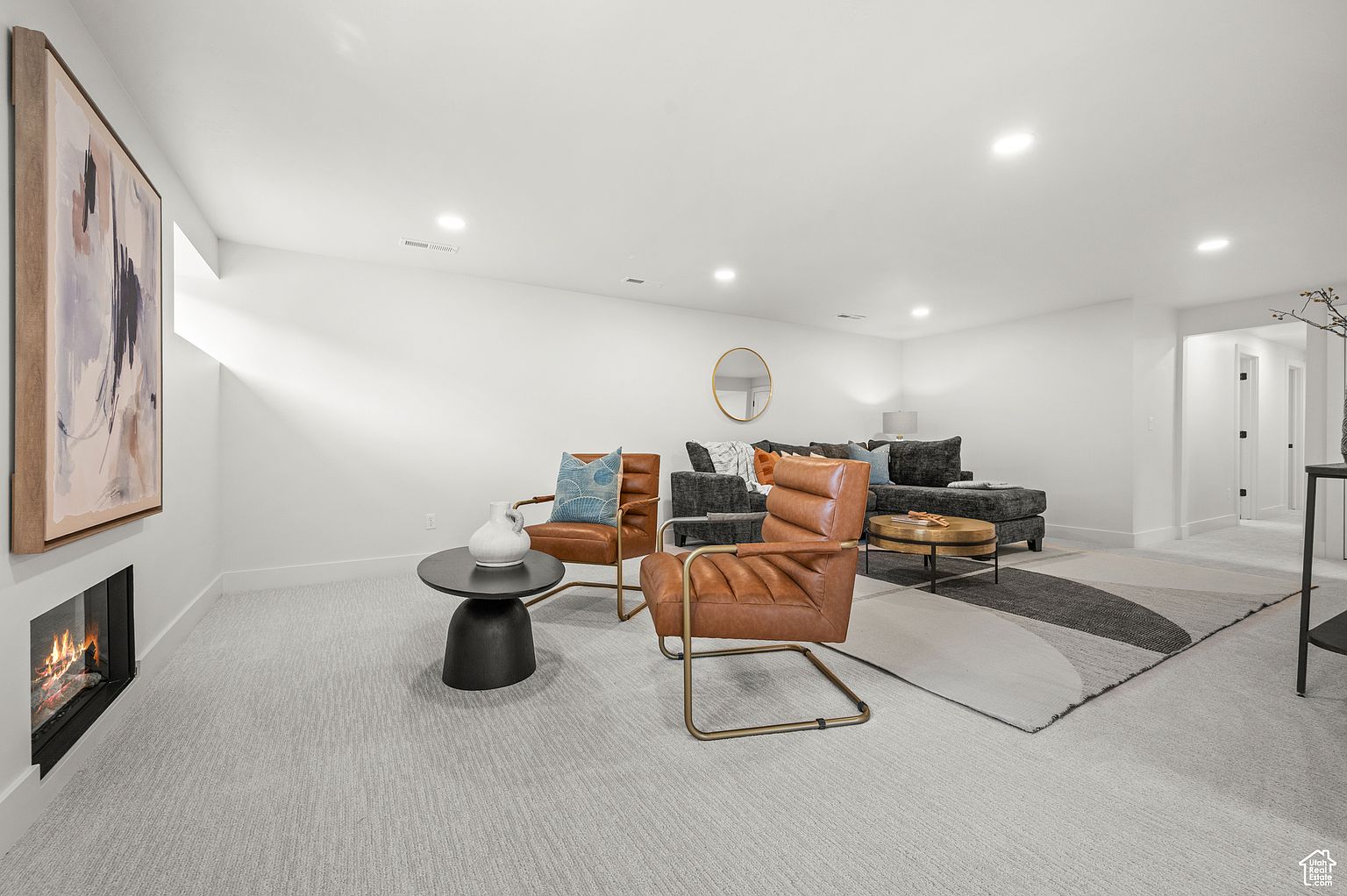 This is an interior shot of a modern living room featuring a gray sectional sofa, two leather chairs, and a round coffee table. A fireplace is built into the wall, and a large abstract painting hangs above it. The room is well-lit with recessed lighting and natural light from a window, creating a bright and inviting atmosphere.