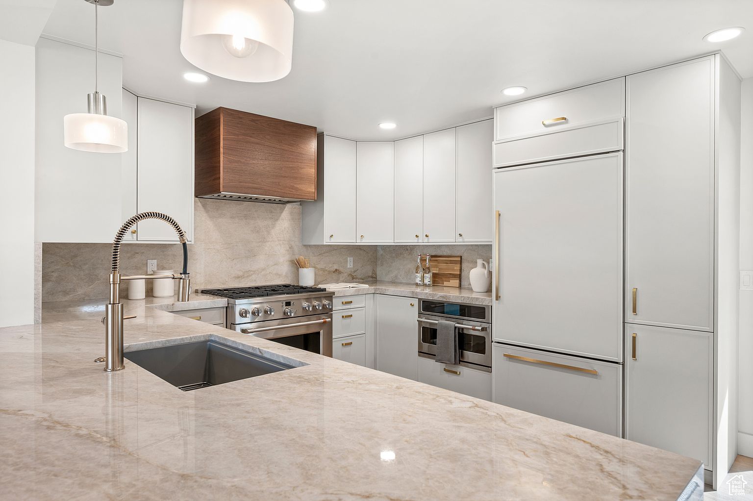 This is a well-lit kitchen featuring white cabinetry with gold hardware, stainless steel appliances, and a large island with a light-colored marble countertop. A modern faucet and sink are prominently displayed on the island, and a wooden range hood adds a touch of warmth to the space. The overall impression is clean, modern, and luxurious.