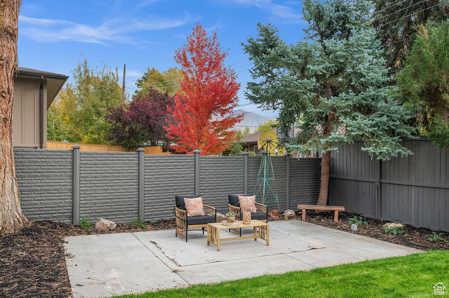This is a backyard patio area featuring a cozy seating arrangement with two wicker chairs and a small bamboo coffee table on a concrete slab. The patio is bordered by a well-maintained lawn, mulch beds, and a combination of gray brick and wood fencing. A vibrant red tree and a blue spruce add color and visual interest to the landscape, creating a serene and inviting outdoor space.
