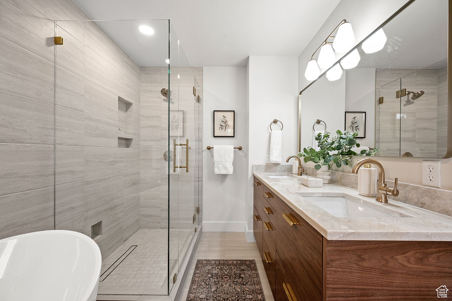 This is a well-lit primary bathroom featuring a glass-enclosed shower with tiled walls and a modern vanity with double sinks and a marble countertop. The bathroom has a contemporary design with gold fixtures and a neutral color palette, creating a luxurious and spa-like atmosphere. A decorative rug adds a touch of warmth to the space.