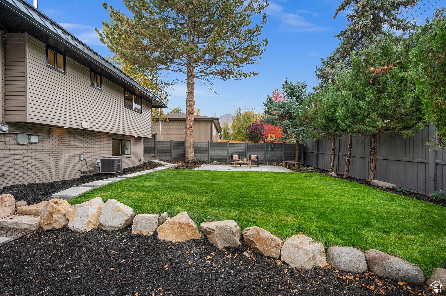 This image showcases a well-maintained backyard featuring a lush green lawn, a concrete patio area with outdoor seating, and a decorative rock border. A dark gray fence provides privacy, while mature trees add shade and visual interest. The overall impression is a serene and inviting outdoor space perfect for relaxation and entertaining.
