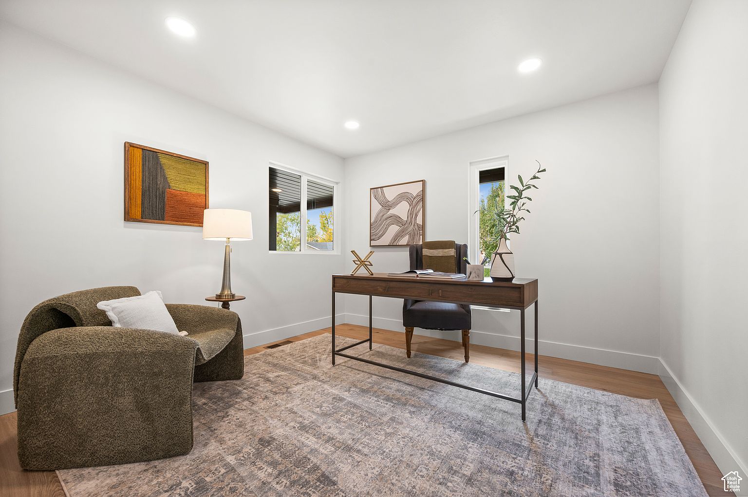 This interior shot showcases a well-lit home office featuring a modern desk with a sleek metal frame, complemented by a comfortable chair. A textured rug anchors the space, while artwork adorns the walls, adding visual interest. The room is bathed in natural light from the windows, creating a bright and inviting workspace.