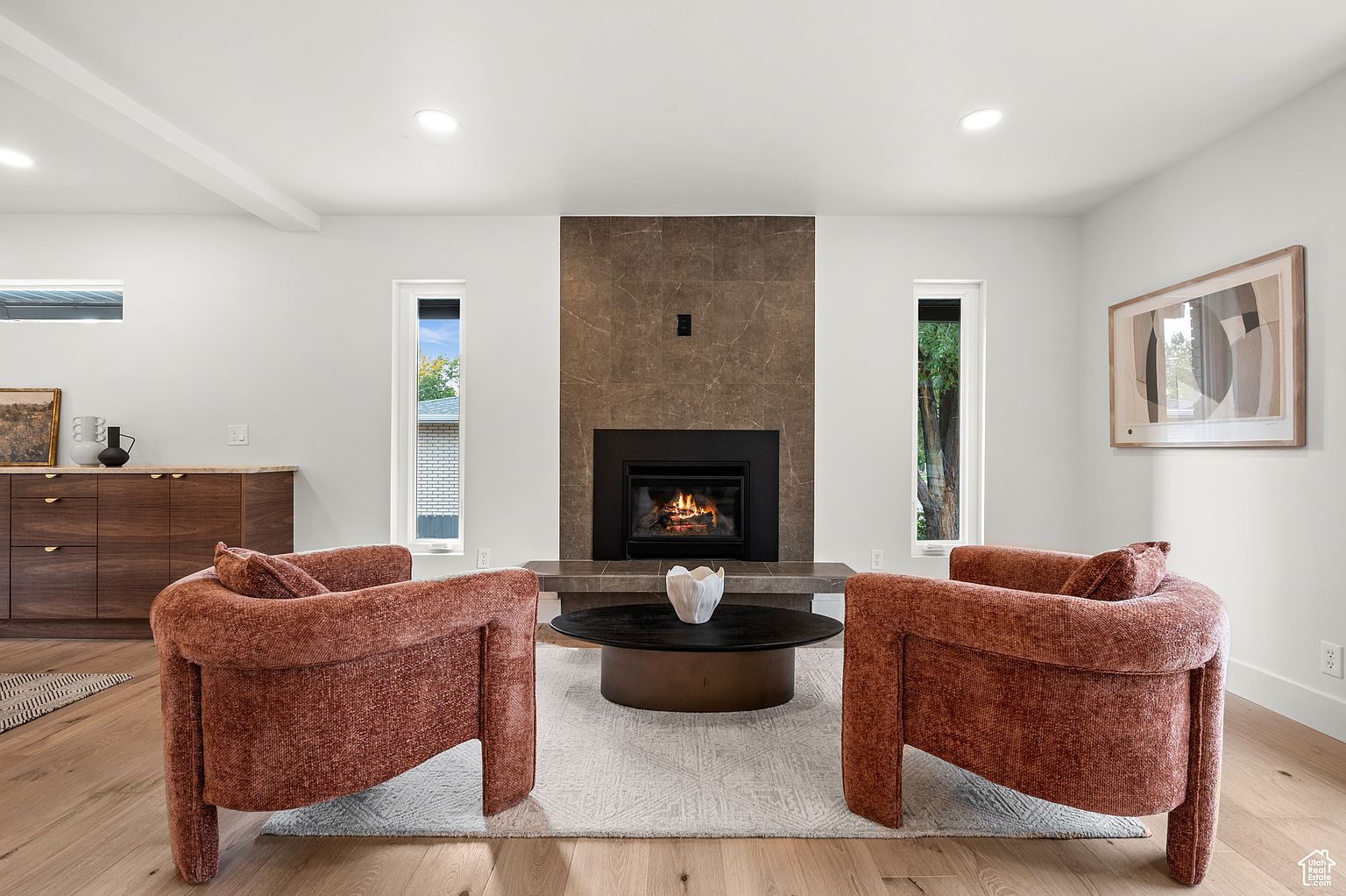 This is an interior shot of a living room featuring two rust-colored armchairs facing each other, flanking a round coffee table with a decorative vase. A modern fireplace with a dark surround and a stone facade is centered between two narrow windows, and a piece of artwork hangs on the right wall. The room has light hardwood floors and white walls, creating a bright and inviting atmosphere.