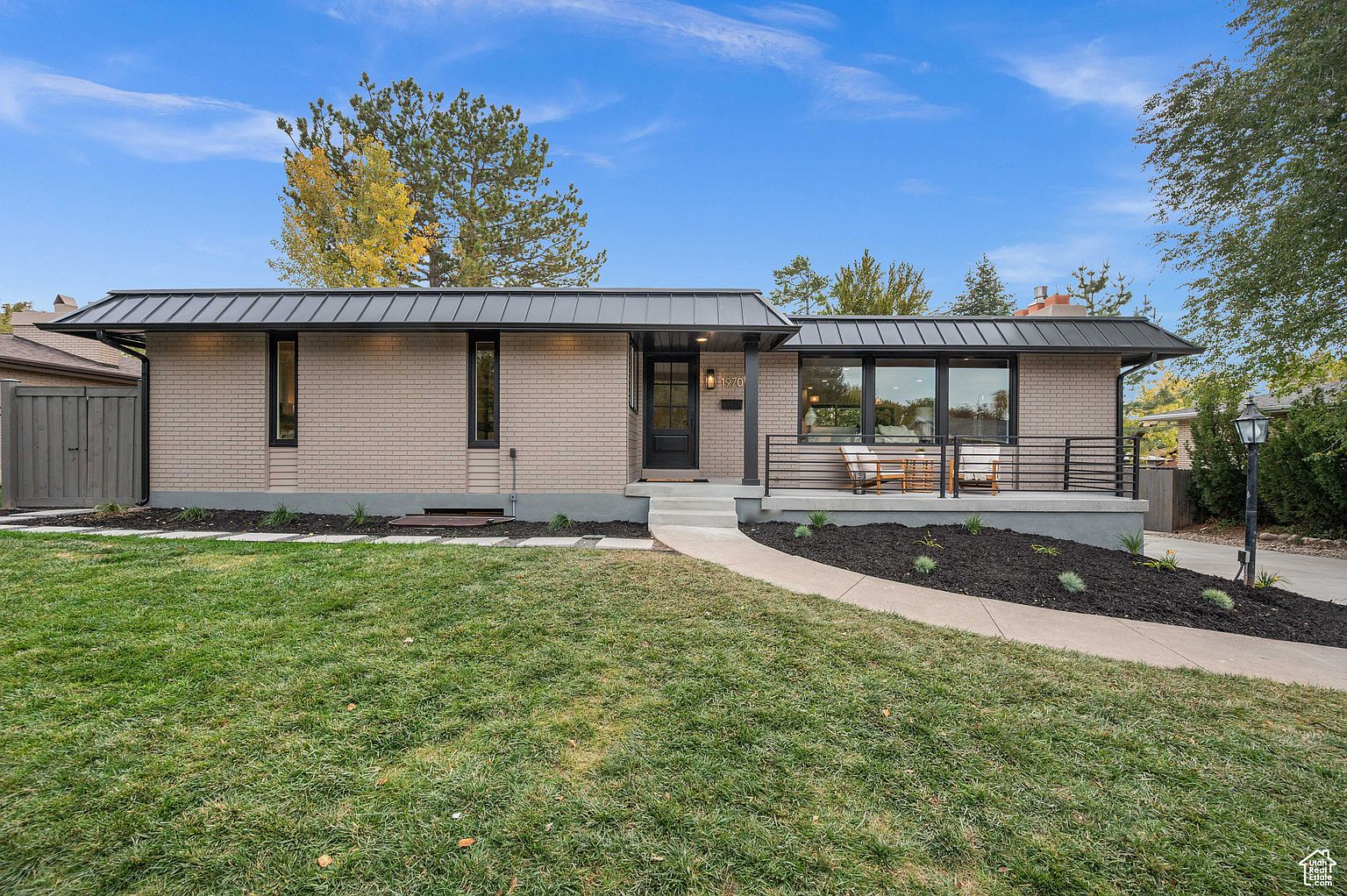 This is a front exterior view of a single-story brick home with a modern design. The house features a dark metal roof, black-framed windows, and a small front porch with a railing. A well-manicured lawn and a concrete walkway lead to the front door, enhancing the property's curb appeal.